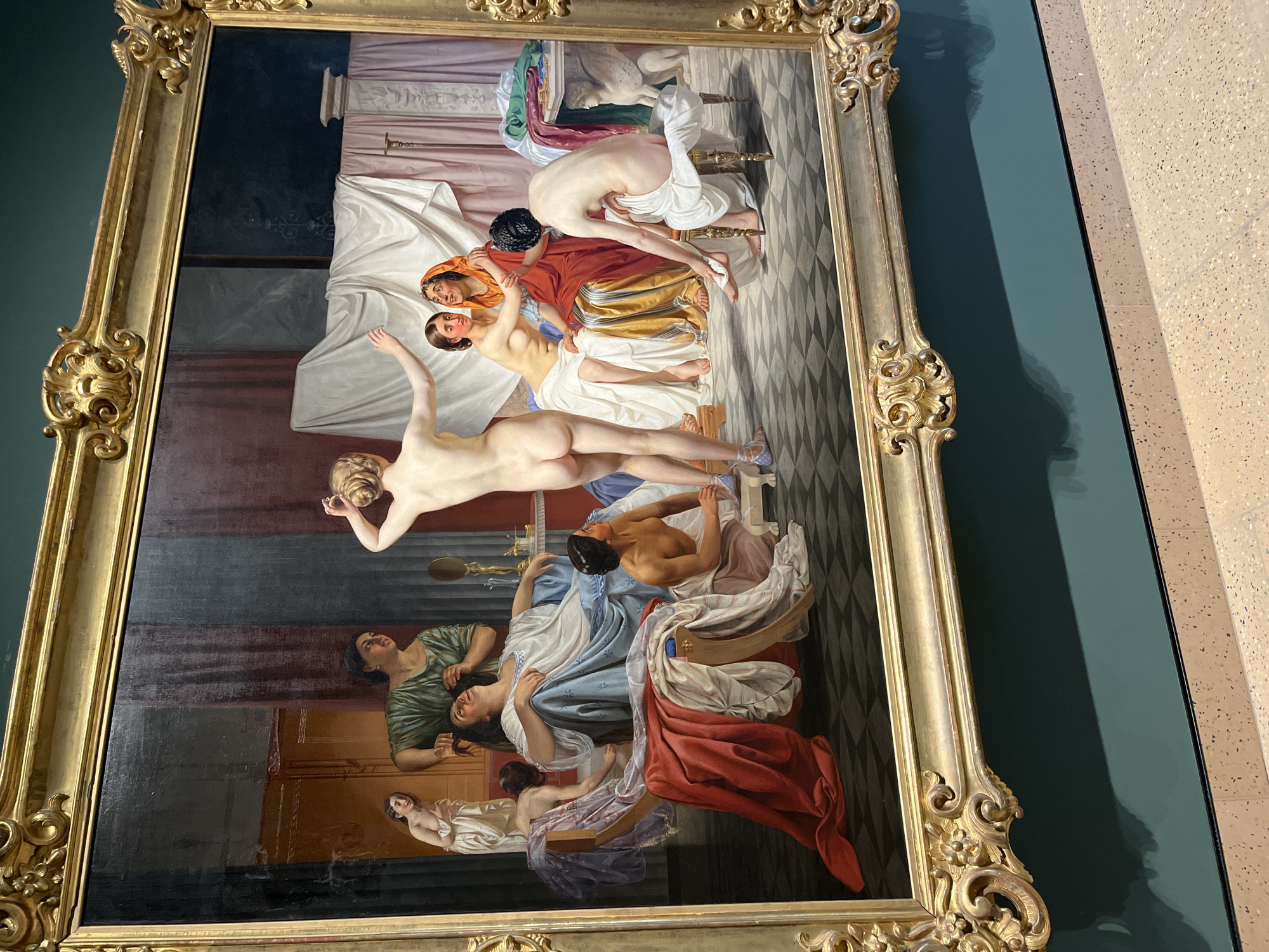 The image depicts a framed painting on a wall, likely in a museum or gallery. The painting itself depicts a historical scene featuring multiple nude or partially clothed figures in a state of apparent distress or turmoil. The figures are arranged on and around a bed, with others present in what appears to be a bedroom or domestic interior. The style of the painting suggests a 19th-century or earlier era. The frame is ornate and gold-colored. The wall behind the painting is a deep teal color, while an adjacent wall has a tan, textured surface. The location is not specified, but the setting is an art institution in an unknown city.