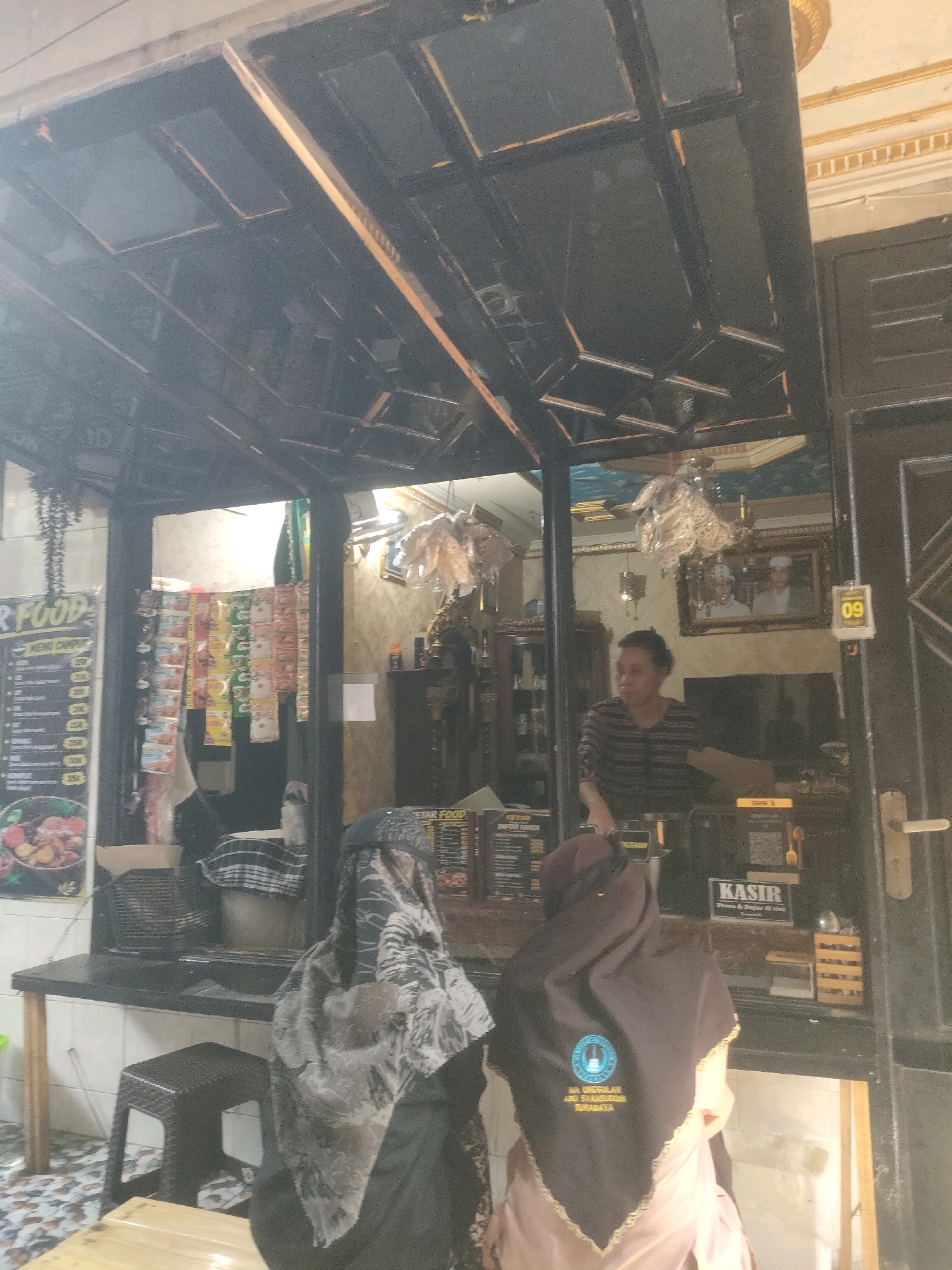 The image depicts an outdoor food stall in Surabaya, Indonesia. A person, likely a vendor, is visible behind the counter, appearing to interact with customers. Two individuals in hijabs are positioned in front of the counter, suggesting they are customers. The stall itself appears to be a small, open-air establishment with a counter, menu boards displaying prices, and shelves stocked with various items. The overall ambiance suggests a casual dining setting.