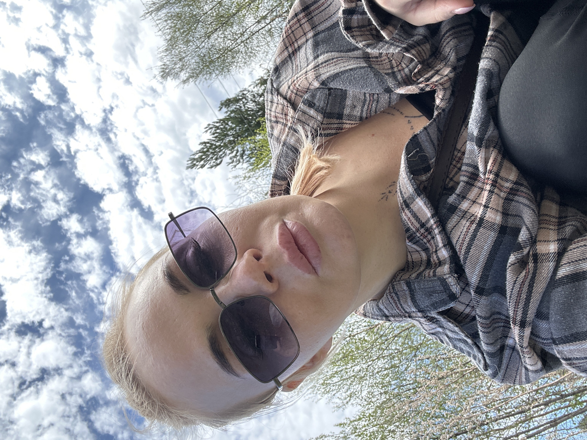A low-angle shot depicts a person wearing sunglasses and a plaid shirt, situated outdoors. The sky, adorned with scattered clouds, is visible in the background, along with foliage suggesting a wooded area. The location is Высокий, Russia. The person is looking down towards the camera.