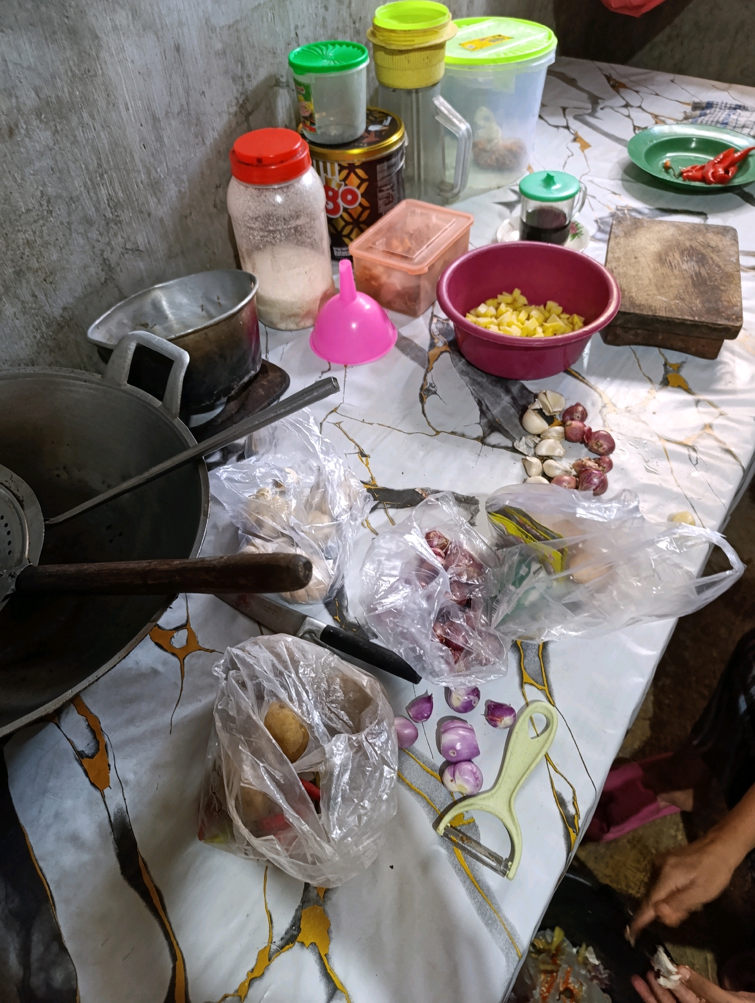 The image depicts a cluttered kitchen counter in what appears to be a home in Sebandung, Indonesia. The scene is a close-up shot focused on various ingredients and cooking utensils, suggesting meal preparation. A large wok is prominent in the foreground, with a metal spatula resting inside. To the right, a chopping board and a green plate with red chilies are visible. Various containers, including a pink bowl filled with chopped yellow vegetables, a jar of salt with a red lid, and several plastic bags containing onions and other produce, are scattered across the patterned countertop. A peeler lies near a pile of sliced shallots and garlic cloves. In the background, a wall with a textured finish and several larger containers are present. The lighting suggests an indoor setting, likely during the day, with no discernable natural light sources. The overall impression is one of a functional, lived-in kitchen where cooking activities are underway.