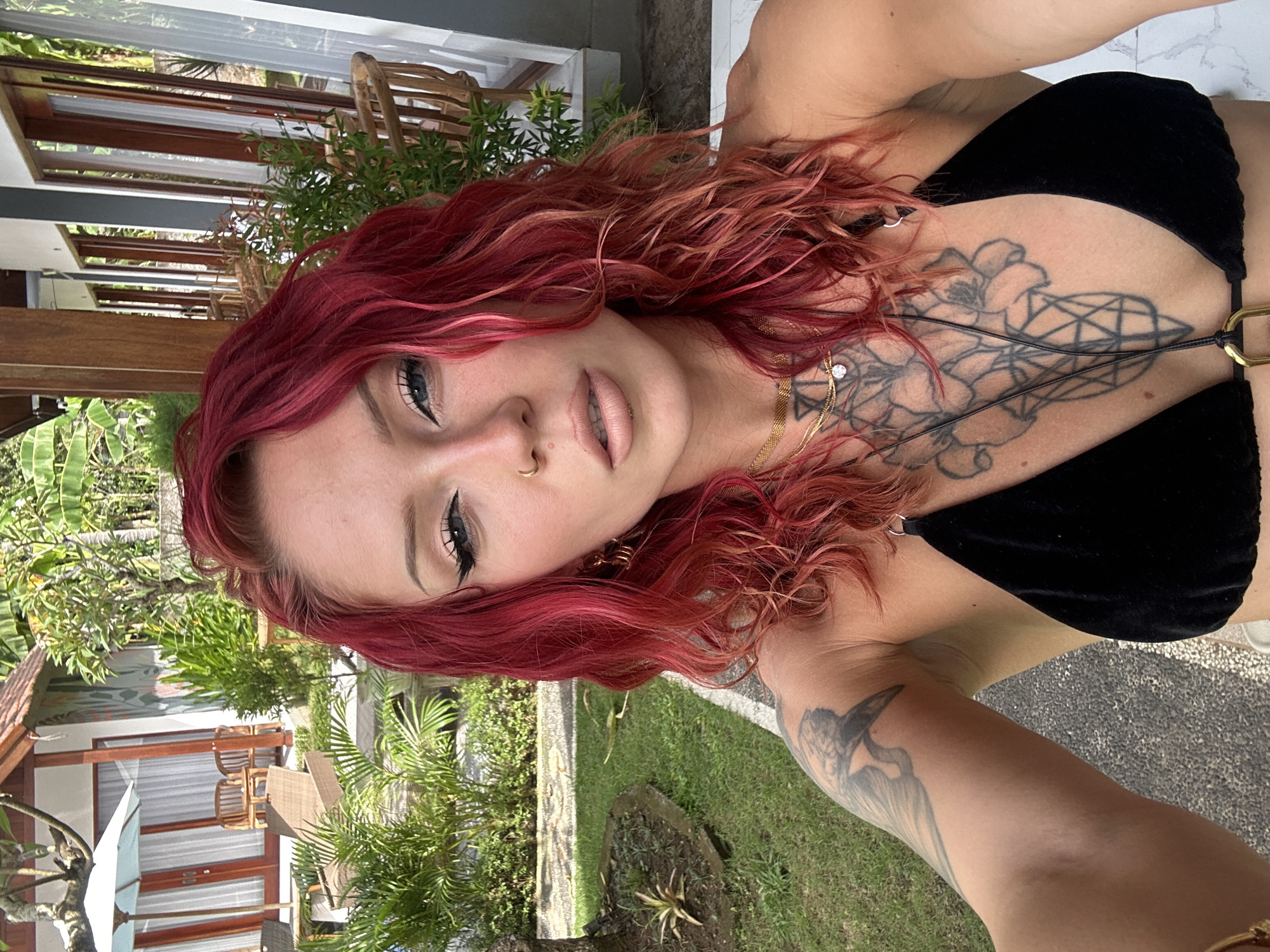 A selfie photograph of a woman with bright red, wavy hair is taken outdoors. She wears a black bikini top, revealing tattoos on her chest and arm. The background shows a tropical garden with green plants and a paved area. Buildings with wooden accents and outdoor furniture are also visible, suggesting a resort or villa setting, possibly in Nusa Penida, Indonesia.