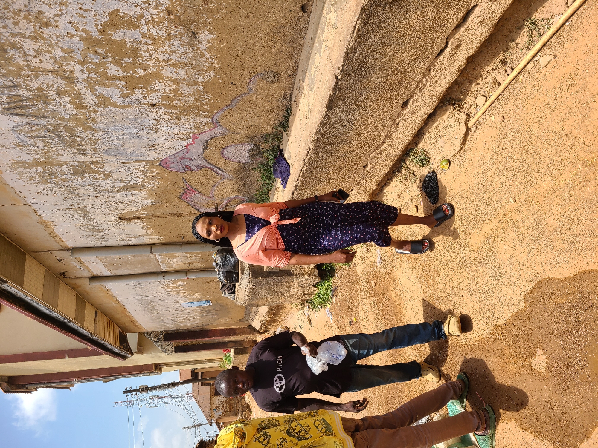 The image depicts an outdoor scene, possibly an alleyway or street in an urban environment. A woman is standing in the alleyway. She is wearing a navy blue dress with white polka dots, and a light pink shirt tied around her waist. She is also wearing black sandals. She has a dark complexion. To the woman's left, a man is standing facing the viewer. He is wearing a black T-shirt with a white logo and blue jeans. He has a dark complexion. The third person in the image is only partially visible wearing a yellow shirt. The ground appears to be dirt. The walls around appear to be concrete.