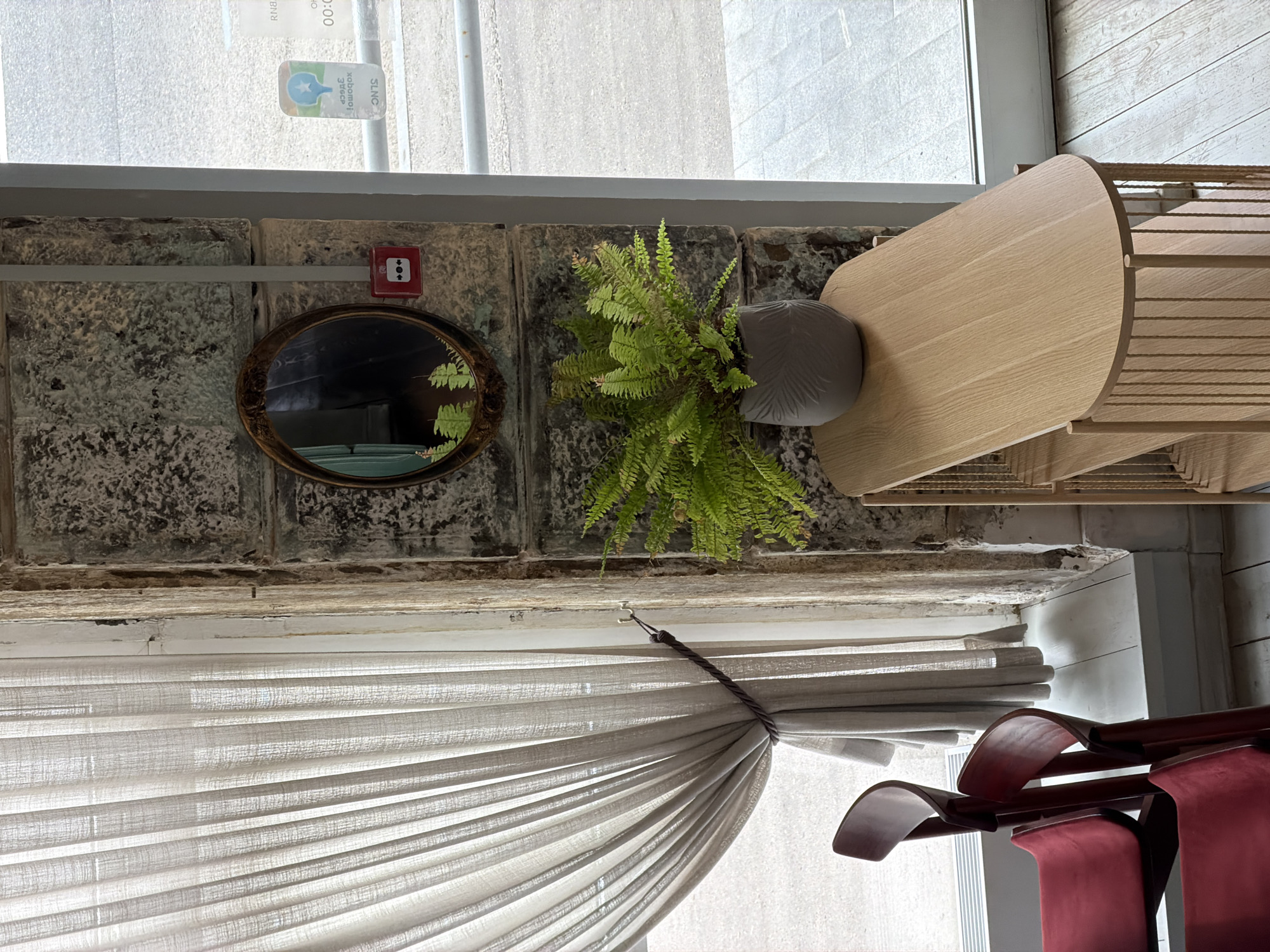 The image depicts a section of an interior wall with a window. The wall appears to be constructed from weathered stone bricks with a gray tone. Above the window is a sign written in Russian with a blue logo on it.
Attached to the wall are a round mirror with a bronze frame and a potted fern. To the right of the fern is a wooden, tiered structure that resembles a display shelf or light fixture.
Below the wall and window, a curtain with horizontal folds is gathered to the side with a dark cord. Partial view of maroon chairs near the curtain.
The image does not provide enough information to confirm the location is Moscow, Russia.