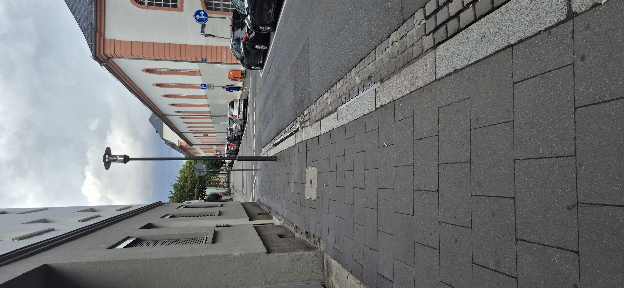 The image shows a street scene in Mainz, Germany, looking down a sidewalk. On the left is a building with a grey facade and horizontal windows. On the right is a long, white building with a pink trim and arched windows. The street is lined with parked cars. A street lamp is visible in the middle ground. A person in a blue outfit is walking on the sidewalk in the distance. The sky is cloudy.