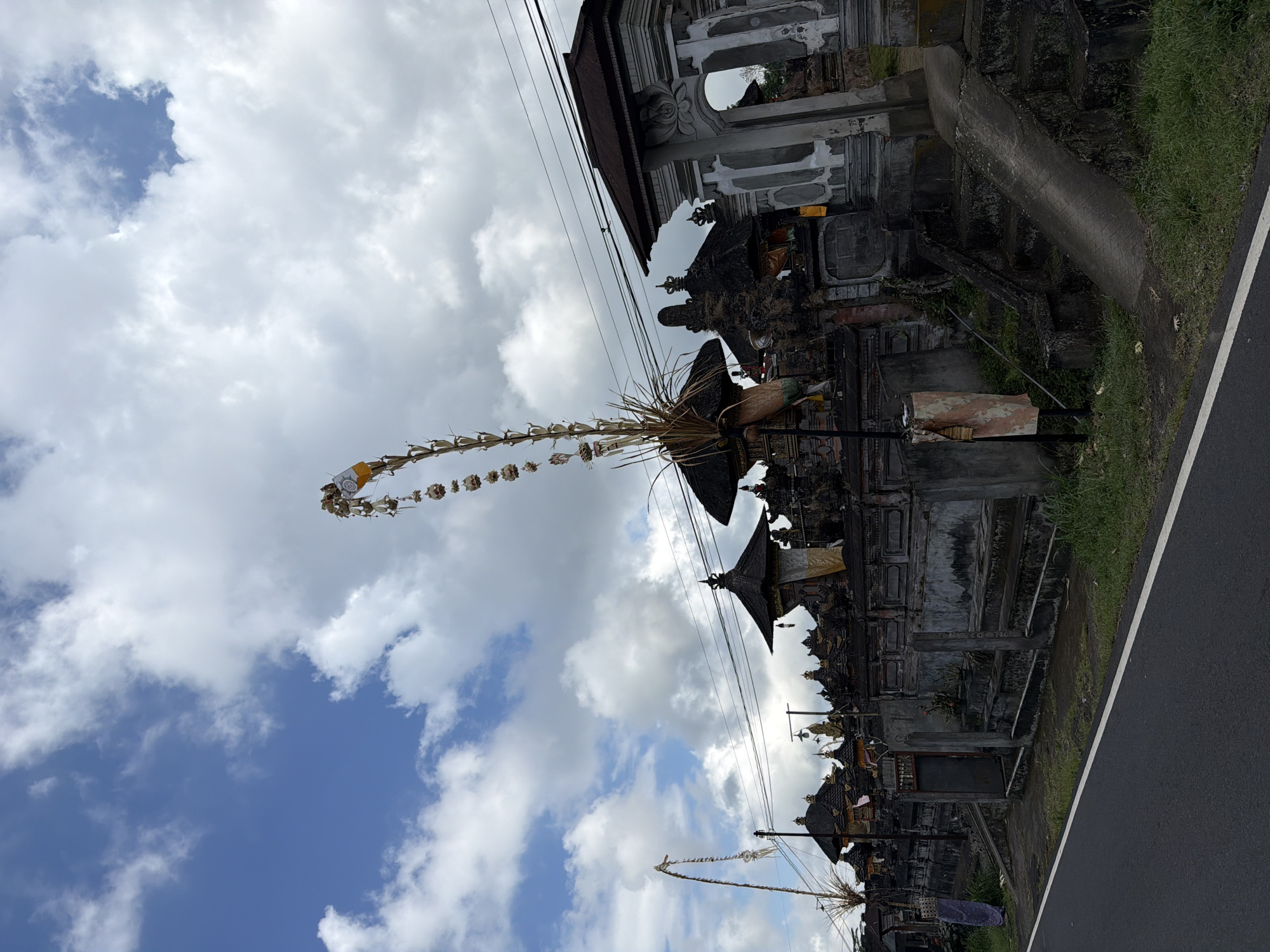 In the city of Susut, Indonesia, a view of a temple complex on the side of a road is captured under a partly cloudy sky. The temple features dark, multi-tiered roofs and elaborate stone walls, with a large, ornate archway visible on the right. Two tall, decorative bamboo poles, known as "Penjor," stand prominently in front of the temple. One Penjor, adorned with offerings and streamers, arches high into the sky on the right side of the frame, while another shorter one is visible on the left. The foreground shows a paved road with a white line marking its edge, and a strip of green grass separates the road from the temple grounds. The overall scene depicts a traditional Balinese temple entrance against a backdrop of a dynamic sky.