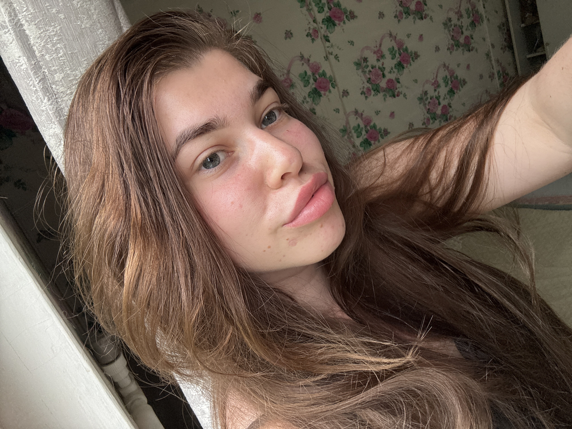 A young woman with long, brown hair and fair skin is captured in a close-up selfie. She has striking blue eyes and full, pink lips. Her skin shows some redness and a few scattered freckles. Her hair is styled loosely, framing her face and falling over her shoulders. The background is a mix of a white, textured curtain on the left and floral wallpaper with pink roses on the right. There's a hint of a white structure in the foreground, possibly a window frame or furniture. The lighting suggests an indoor setting, potentially a bedroom or living area. The image is a casual portrait, likely taken at home in Мещёры, Russia, given the context.