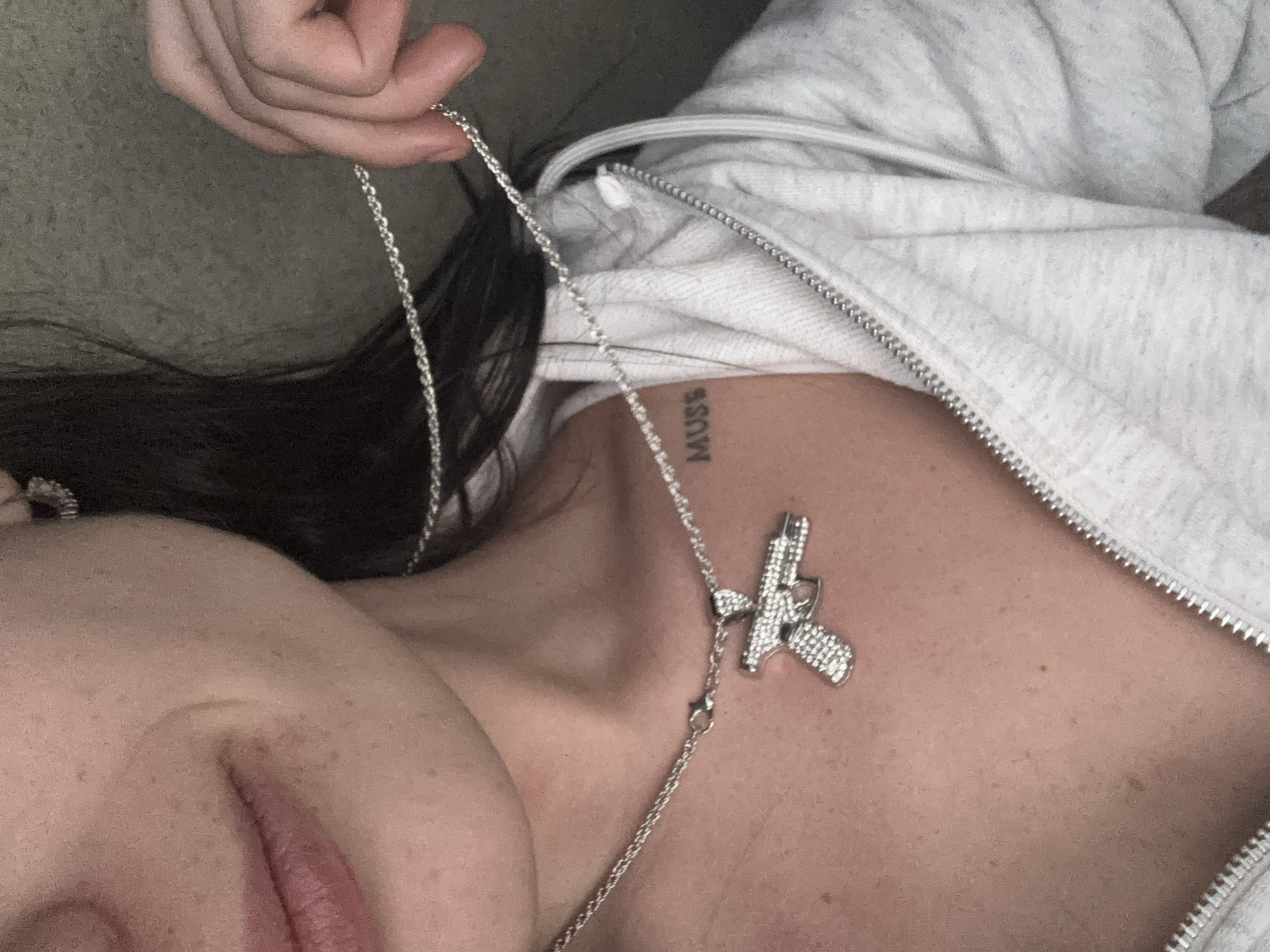 A close-up shot features a person with dark hair and fair skin wearing a light gray zip-up hoodie. They are holding a silver chain necklace with a gun pendant, which lies on their chest. A tattoo reading "MUSS" is visible on their collarbone. The person's face is partially visible, focusing on their lips. The background appears to be a neutral-toned surface. There is no indication of a location in Buenos Aires, Argentina.