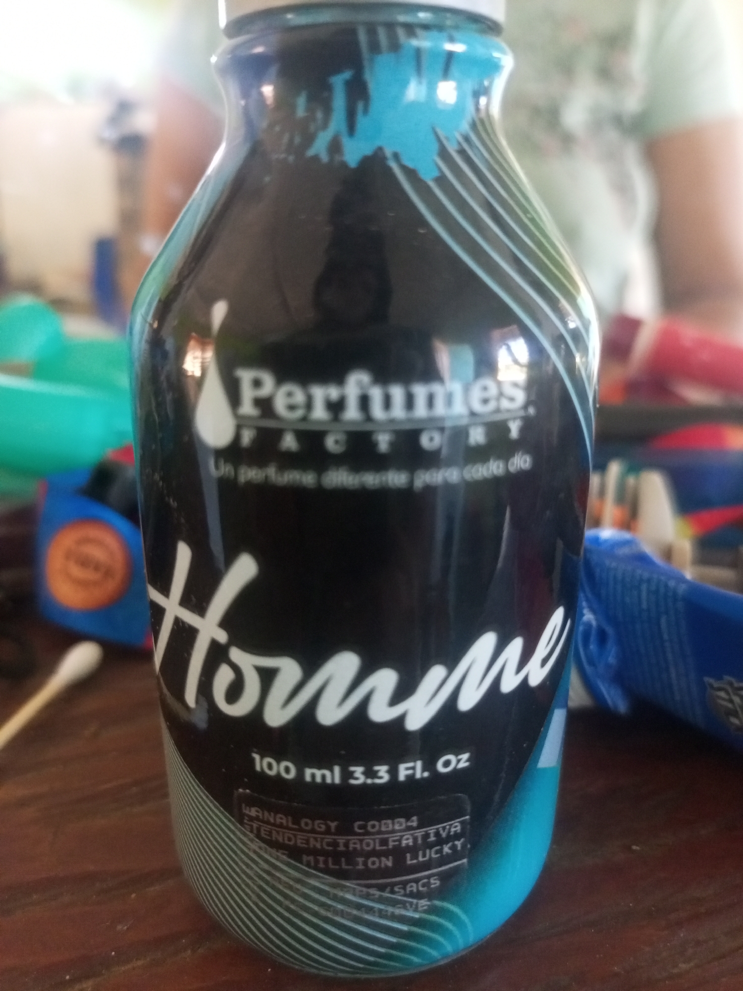 This image is a close-up, eye-level shot of a bottle of "Perfumes Factory" brand cologne for men, labeled "Homme." The bottle is dark with teal and white accents, featuring a stylized wave design. Text on the bottle indicates it contains "100 ml 3.3 Fl. Oz." and lists some ingredient information. In the background, a person is partially visible wearing a light-colored shirt. The environment appears to be indoors, with various out-of-focus objects suggesting a domestic or retail setting. The specific location in La Tagua, Venezuela, cannot be determined from the visual information provided in the image.