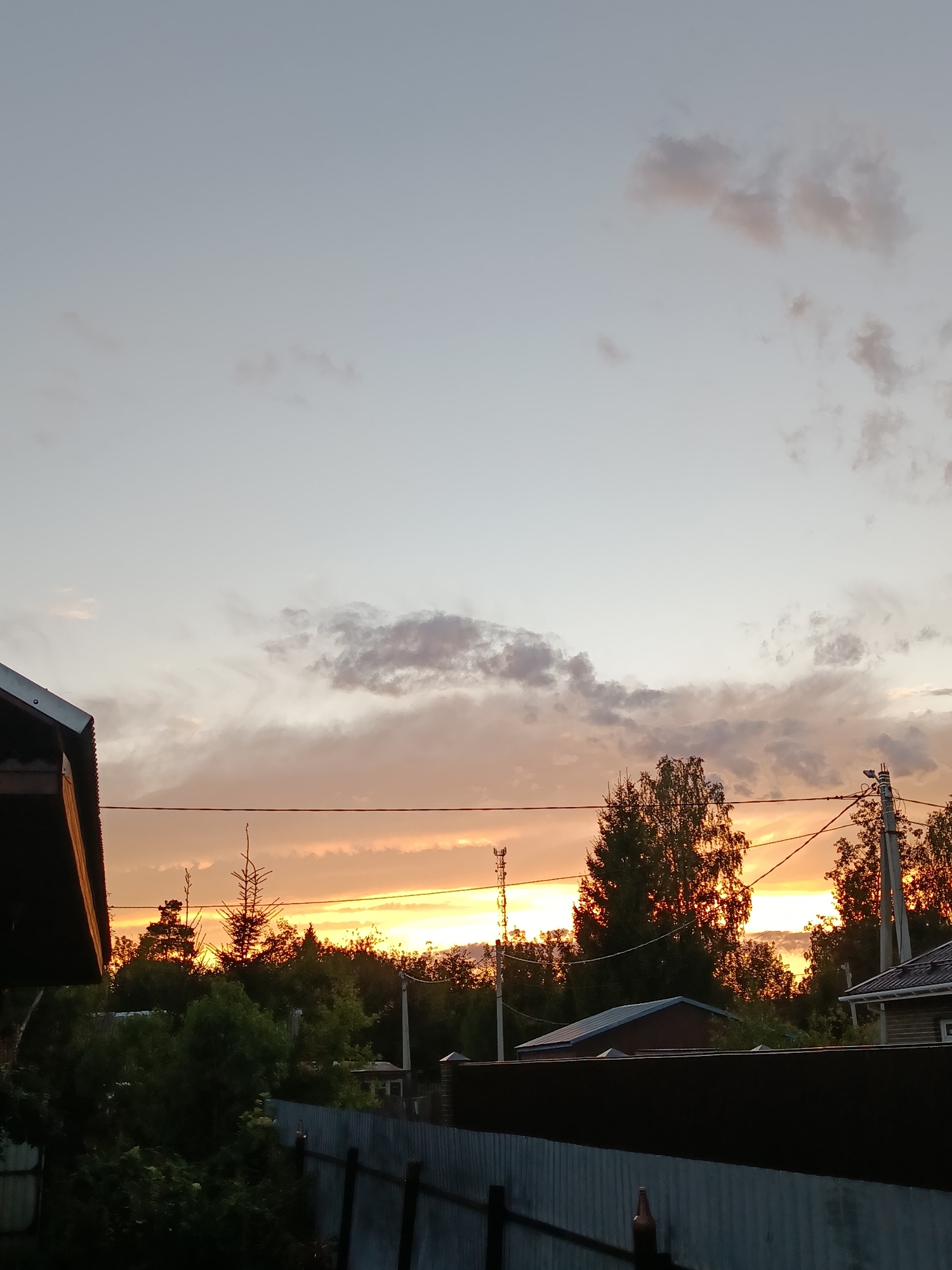 The image depicts a rural landscape at dusk, likely in or around Vereya, Russia. The sky is a gradient of soft blues and grays, transitioning into warm orange and yellow hues near the horizon, indicating the setting sun. Clouds are scattered across the sky, with some appearing wispy and others more defined and grey.
In the foreground, the dark, sloped roof of a building is visible on the left side. A metal fence runs horizontally across the lower portion of the image, with some vegetation and trees peeking over it. Beyond the fence, there is a dense line of trees silhouetted against the bright sunset.
Several man-made structures are present. On the right side, a house with a dark roof and a section of white siding is visible. A power pole with electrical wires stretches across the scene, connecting to another pole on the far right. A smaller utility tower or antenna is also visible in the distance on the left side.
The environment appears to be a residential area with natural elements, suggesting a dacha or village setting. There are no people visible in the image, and no specific actions or interactions are captured. The overall mood is tranquil, characteristic of the end of a day.