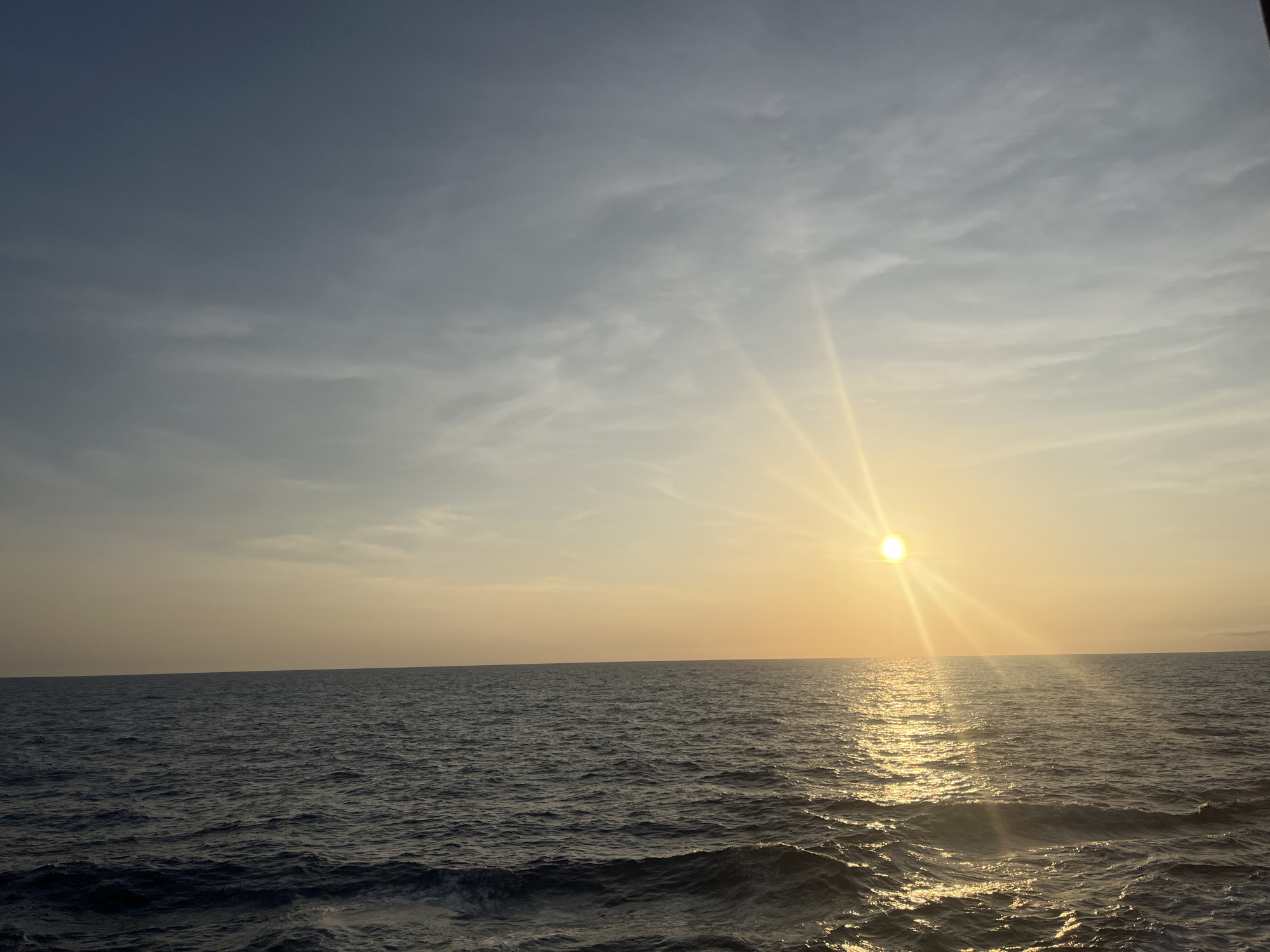 The image shows a body of water, possibly an ocean, with the sun setting on the horizon. The sky is a mix of blue and yellow hues with some clouds. The water reflects the sunlight, creating a shimmering effect. The location appears to be a coastal area, but the specific city is unknown.