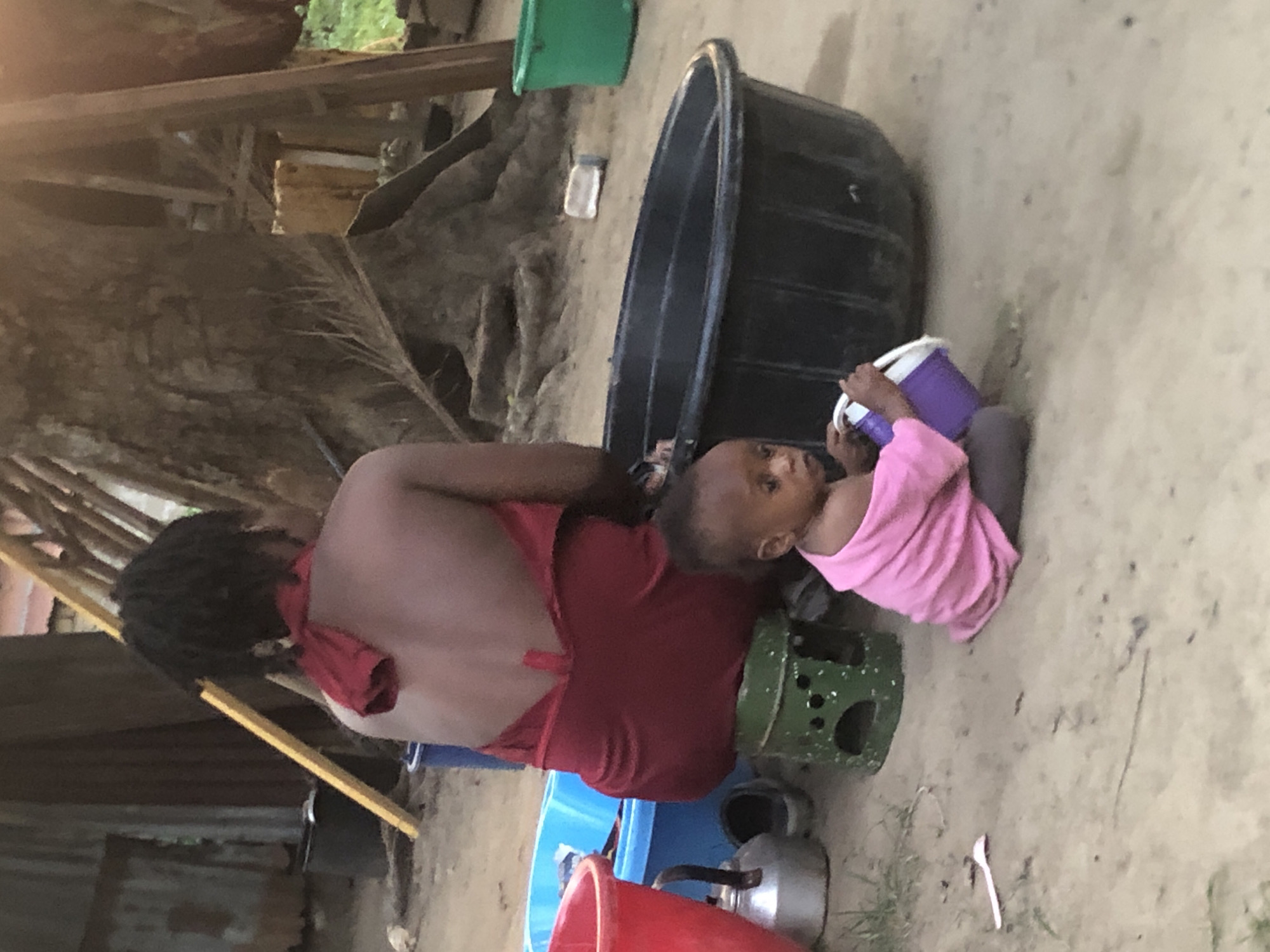 The image shows a woman sitting outside on a blue plastic stool. She has her back to the camera and is wearing a red dress. Beside her is a child wearing a pink shirt, lying on their back, and holding a purple cup. Next to the woman is a green metal container with holes in it. Behind the woman are various containers, including a red bucket, a blue basin, and a black tub. There is a kettle on the ground, and a spoon nearby. The background consists of a simple structure with wooden supports. The location is presumed to be Atani, Nigeria.