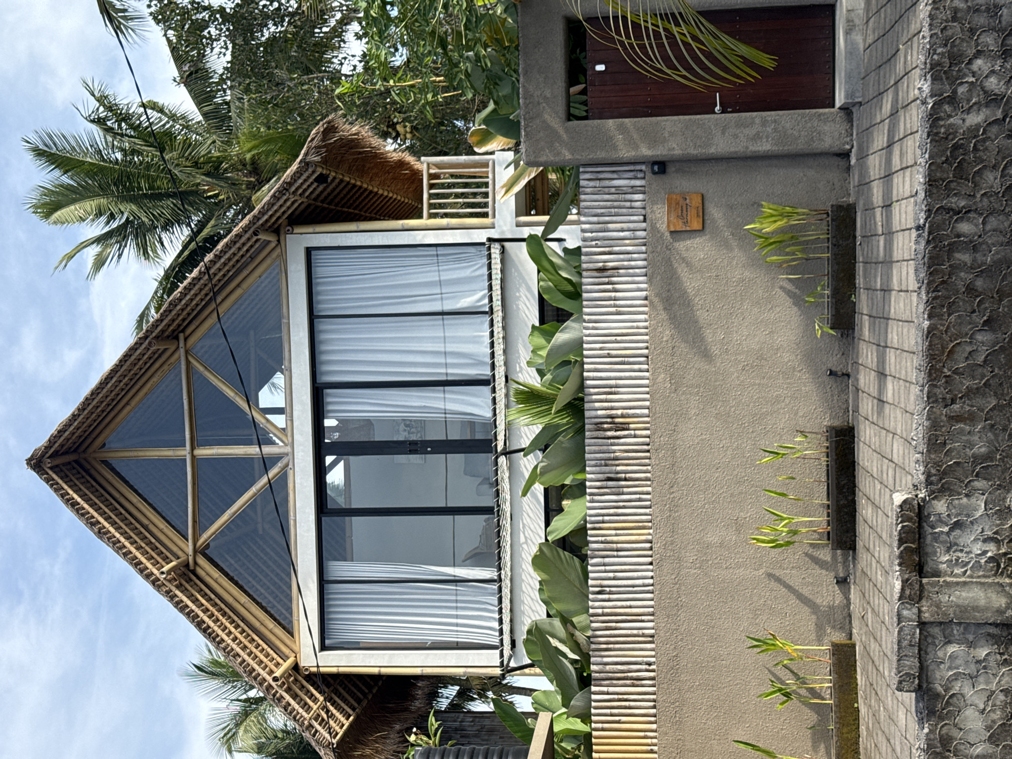This media file captures a modern building in Tampaksiring, Indonesia, showcasing a blend of contemporary design with traditional tropical elements. The building features a prominent, steeply pitched roof with a triangular glass section framed by bamboo, topped with a thatched roof. Below this, large, multi-paneled glass windows are visible, covered internally by white blinds or curtains.
The building's facade is primarily composed of textured concrete, with a decorative section adorned with horizontal bamboo slats. Adjacent to the windows, large-leafed green plants contribute to a lush, integrated natural aesthetic. Further along the wall, several vertical concrete planters are embedded, each containing slender green plants that cast distinct shadows, indicating bright daylight.
In the background and extending above the building, large palm trees with green fronds are visible against a clear, light blue sky with scattered clouds. A dark wooden door is set into a concrete structure to the right of the main building section.
A small, rectangular wooden sign is affixed to the textured concrete wall. Upon closer inspection, it reads "The Green Terrace" on the top line, followed by "Tempatku" (meaning "my place" in Indonesian) on the second line, alongside a small, faint graphic.
No direct human activity is depicted; the scene focuses on the architecture and its natural surroundings. The bright lighting and clear shadows suggest it is daytime, likely morning or afternoon, under pleasant weather conditions. The overall impression is one of a tranquil, tropical dwelling designed to integrate with its environment.
