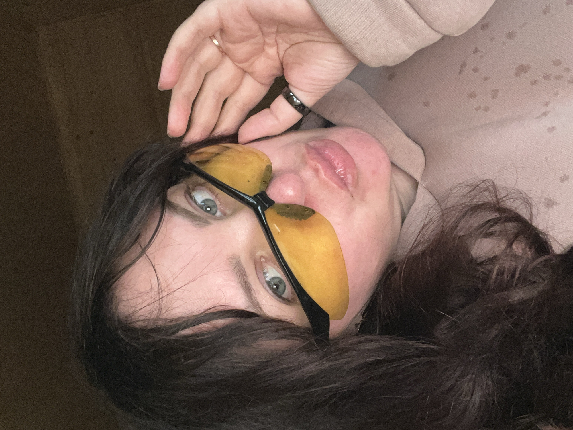 This close-up portrait, captured in Protasy, Russia, features an adult female posing for the camera. Her face is angled upwards and tilted, with her gaze directed forward. She has dark hair, light-colored eyes (likely blue or green), and full, glossy lips that are slightly parted.
She is wearing distinctive glasses with dark frames and unconventional yellow-orange, semi-translucent lenses that appear to have a textured or irregular surface. Her left hand is raised towards her head, with a thin, gold-toned ring on her ring finger and a thicker, dark-colored ring on her middle finger. She is dressed in a light-colored, possibly beige or pale pink, hooded sweatshirt or sweater, which shows several scattered dark spots or stains on its fabric.
The background is a dark, featureless surface, suggesting an indoor or confined setting. The lighting illuminates her face, highlighting her features. Her expression is composed and direct, indicating a posed shot. There is no visible text in the image.