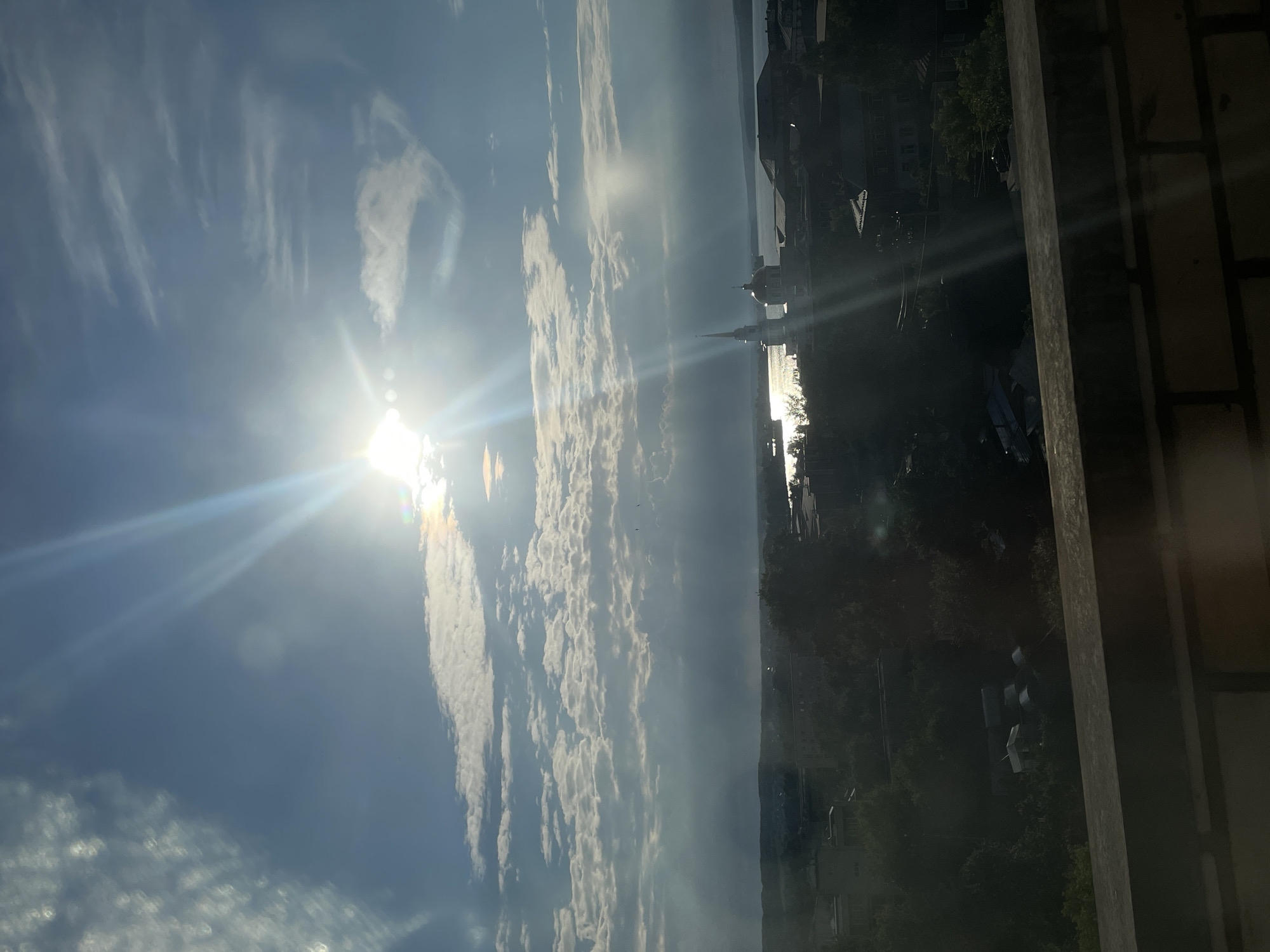 The image shows a cityscape of Votkinsk, Russia, viewed from an elevated perspective, possibly a rooftop. The sun is prominently positioned in the upper right quadrant, casting strong rays and lens flare across the sky. Wispy clouds are scattered throughout the bright blue sky. In the mid-ground, a body of water, likely the Votkinsk Reservoir, is visible. On the shore, a white building with a tall spire, characteristic of a church or cathedral, stands out. Several other buildings with red roofs are visible throughout the cityscape. Dense green trees partially obscure some of the buildings in the foreground and mid-ground. The foreground is dominated by a brick ledge. The overall scene depicts a sunny day in a Russian city.
