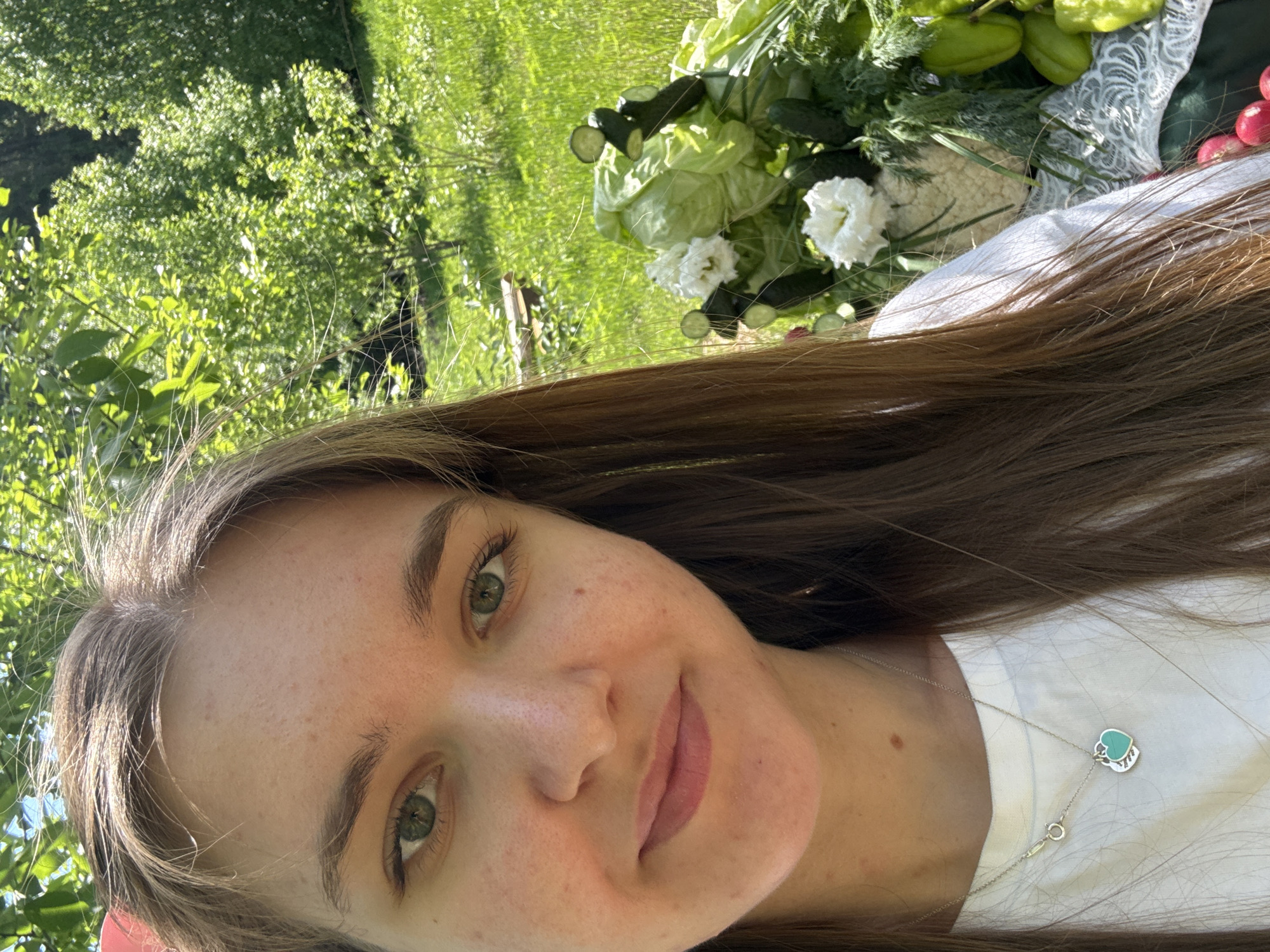 The image shows a selfie of a young woman with light skin, green eyes, and brown hair. She is wearing a white shirt and a silver necklace with a heart-shaped pendant. Behind her, there's a bouquet of vegetables and white flowers, including cucumbers, radishes, and lettuce. The background is a green, grassy area with trees, suggestive of a park or garden. There is no verifiable data to confirm a specific location like the city of Sibir' in Russia. The scene is bathed in natural sunlight, indicating it is daytime. The woman is smiling slightly, looking directly at the camera.