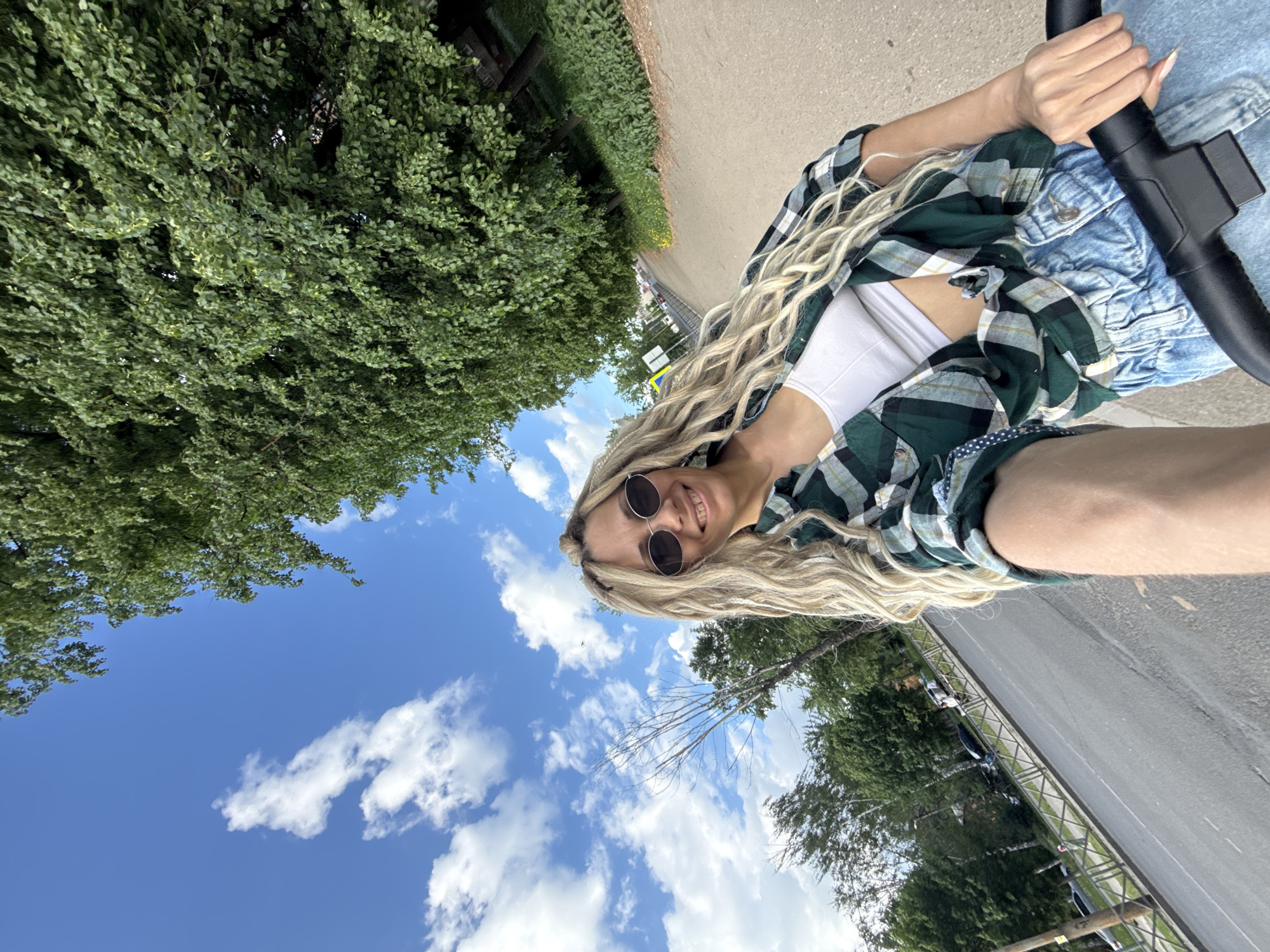 A blonde woman with sunglasses is holding a black baby stroller while standing on a sidewalk next to a road. The view is from her perspective, angled upwards. She is wearing a green and black plaid shirt, a white top, and denim shorts. The sky is blue with scattered white clouds. Green trees line the road. The location is Rusanovo, Russia, based on the availability of images from this location.