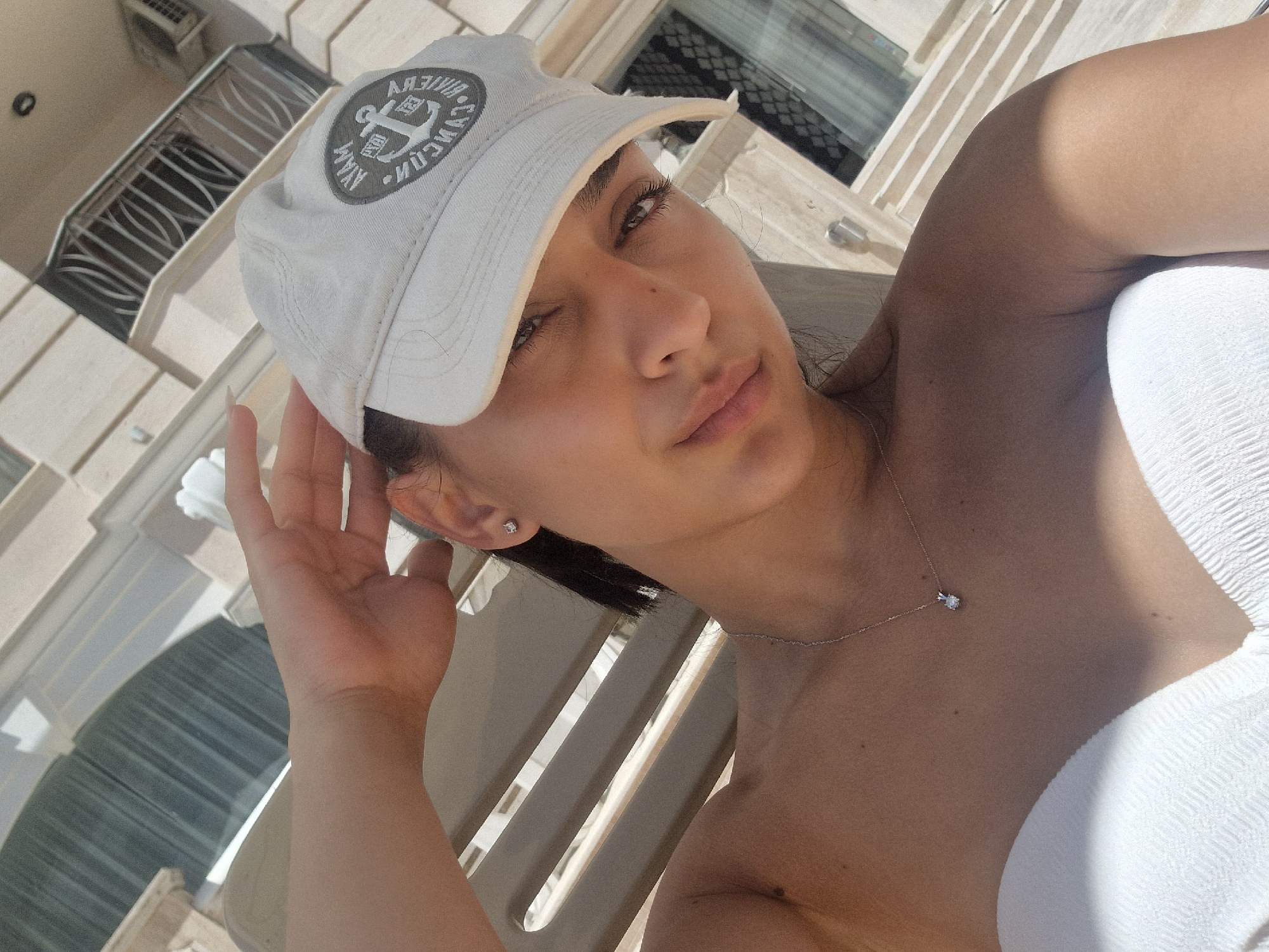 A young woman with dark hair, wearing a white baseball cap and a white strapless top, is positioned in the foreground of the image. She is looking towards the camera with her right hand touching the brim of her cap. A diamond pendant necklace is visible on her neck. The background consists of a building with ornate architectural features and what appears to be a balcony with railings. Sunlight casts shadows on the woman and her surroundings, suggesting an outdoor setting, possibly a balcony or patio.