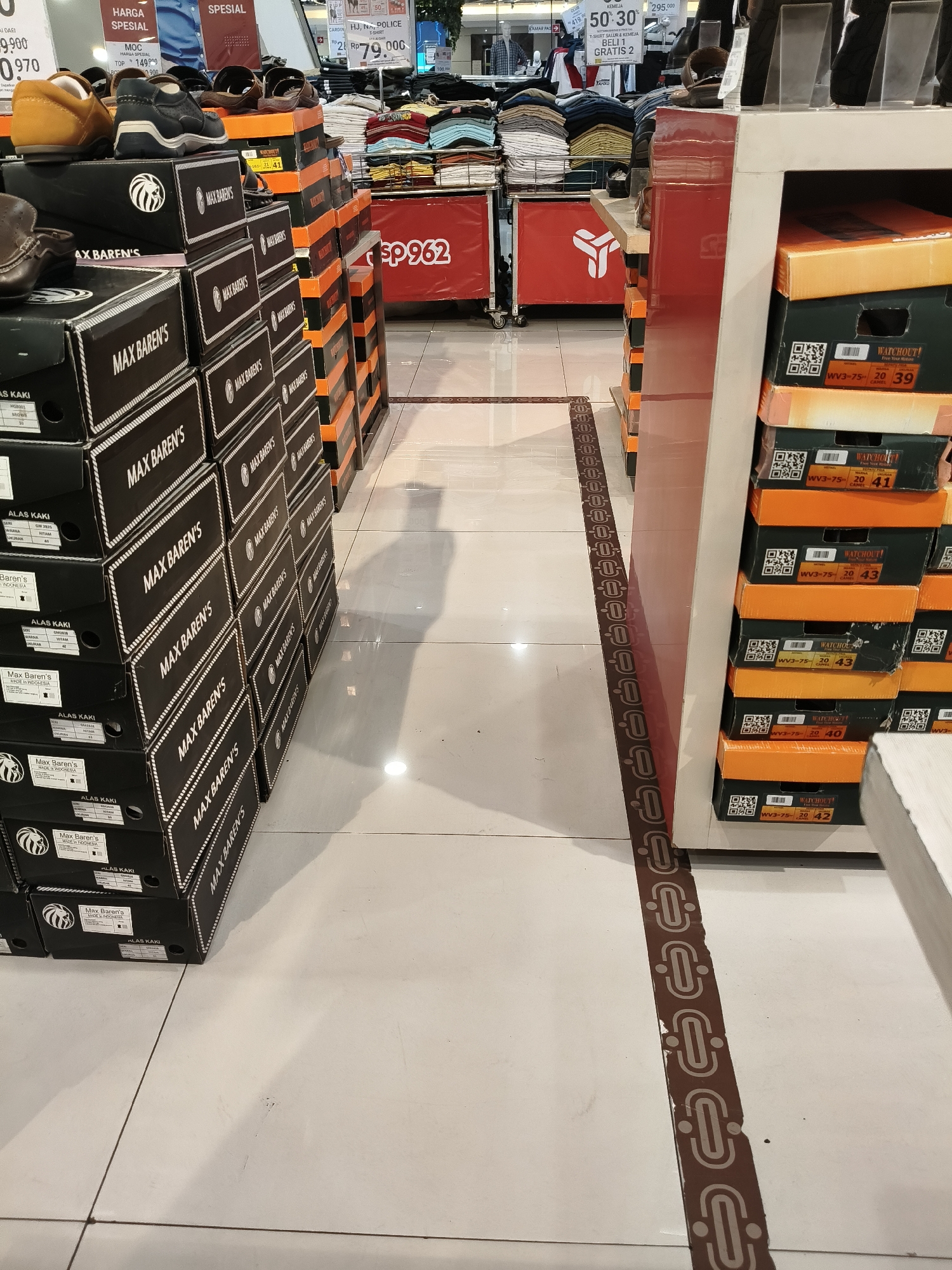 The image depicts an indoor retail environment, likely a shoe store, characterized by stacks of shoe boxes and clothing displays. The flooring is tiled, and a decorative brown strip with an oval pattern runs along the right side of the image, separating the display area from the walkway.
In the foreground, a substantial collection of black shoe boxes with white lettering reading "MAX BAREN'S" is stacked high. Some of these boxes have labels indicating shoe details. Partially visible on top of some boxes are a few pairs of brown and black leather shoes.
To the right, another stack of shoe boxes is visible, these are predominantly orange and green. Labels on these boxes include brand names like "WATCHOUT!" and size information such as "40", "41", "42", and "43".
In the mid-ground and background, clothing items are displayed on racks. One rack prominently features stacks of folded t-shirts in various colors. Further back, signs with Indonesian text and prices are visible, suggesting this is a commercial establishment in Indonesia. One sign displays "HARGA SPESIAL" (Special Price) and "SPESIAL". Another sign reads "ep 962". The presence of Indonesian language on the signs strongly suggests the location is in Indonesia. The specific mention of "Bandung City, Indonesia" in the prompt leads to the inference that this scene is within a retail store located in that city, although no specific landmarks within Bandung City are depicted in the image itself.
No individuals are clearly visible or interacting in the scene. The focus is on the product display and the store environment. The lighting suggests a well-lit indoor space, with reflections on the tiled floor indicating overhead illumination.