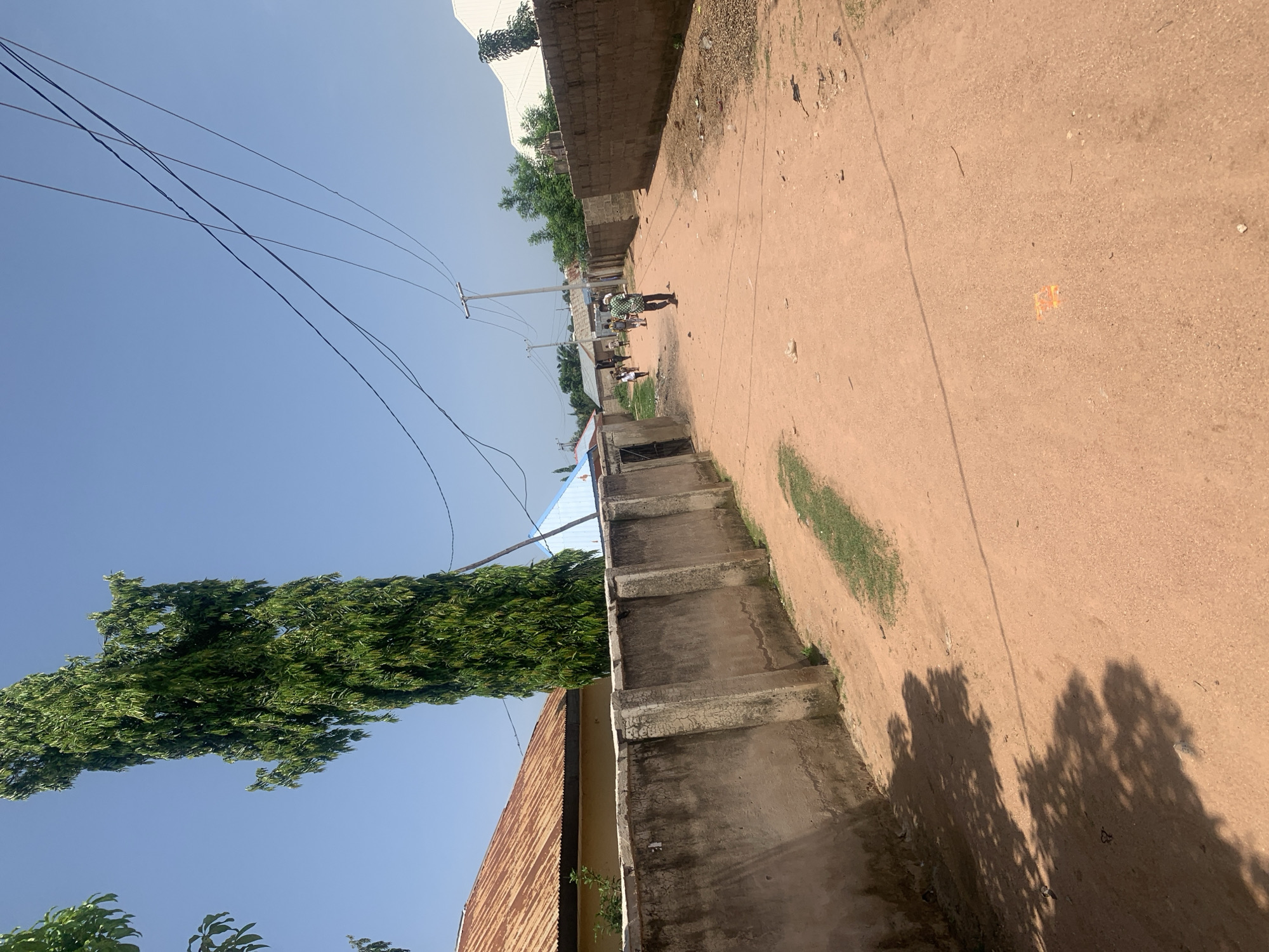 The image depicts a dusty, unpaved street in Jalingo, Nigeria. The street is lined with a low, concrete wall on the left, behind which a house with a corrugated metal roof is visible. Further down the street, a tall, slender tree with dense green foliage stands prominently against a clear blue sky. Power lines are strung overhead.
On the right side of the street, a brick wall separates the road from a property. The road itself is a reddish-brown dirt, showing signs of wear and tear, with scattered debris. Several people are walking or standing in the distance, suggesting a populated area. One individual in the middle ground is walking away from the camera, wearing a patterned shirt. The scene appears to be under bright sunlight, casting shadows from trees onto the road. In the far distance, various buildings and vegetation are visible, indicating a semi-urban or suburban environment.