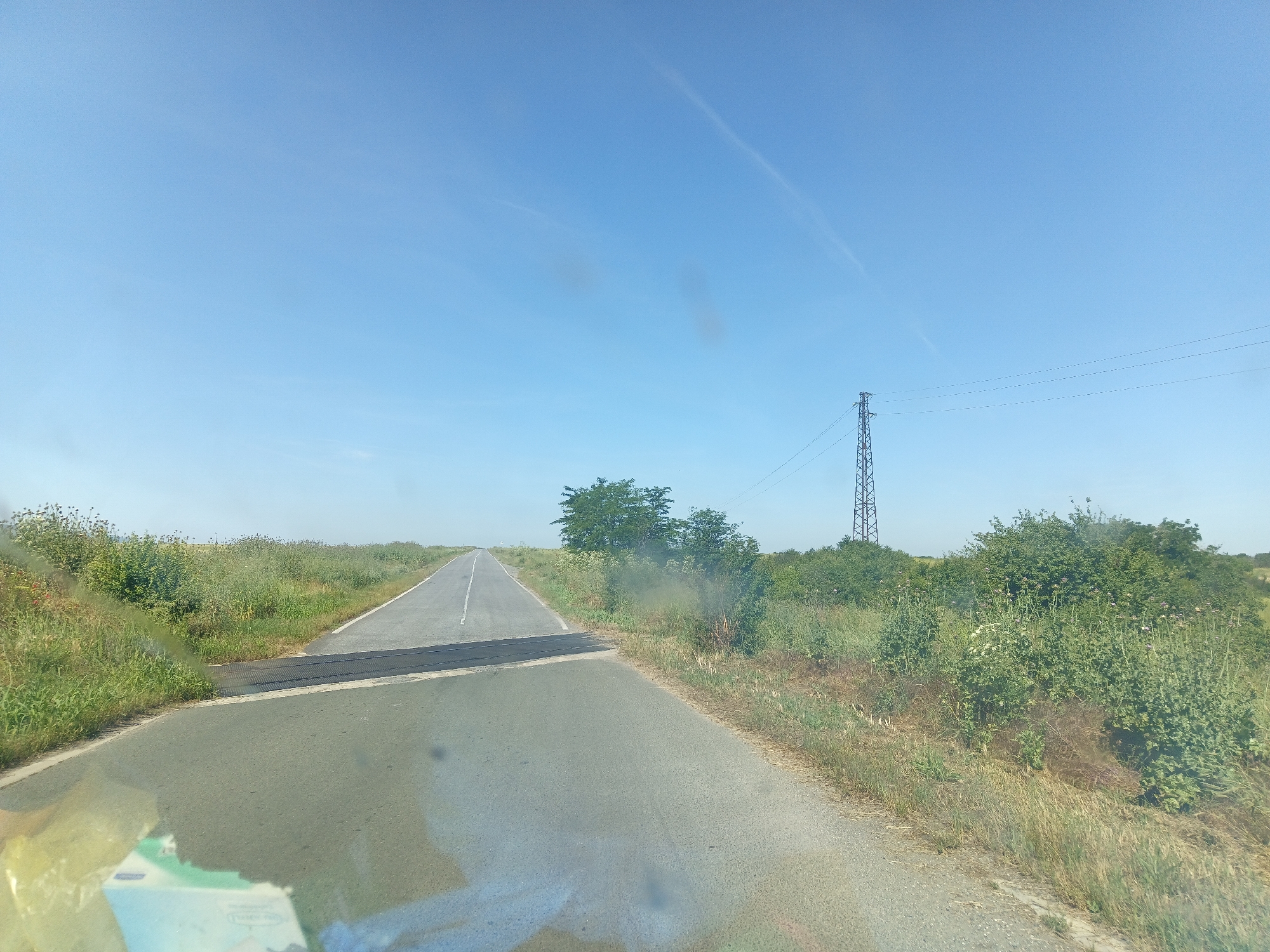 This is a wide-angle, eye-level, exterior shot taken from a moving vehicle on a paved road in General Inzovo, Bulgaria. The sky is clear and blue with a few faint contrails. A lone electricity pylon stands tall on the right side of the frame, with power lines stretching across the sky. The road ahead is empty and stretches into the distance, bordered by green vegetation and fields. The foreground shows the road surface and the vehicle's dashboard, which has some smudges. The overall scene depicts a rural landscape under a bright, sunny sky.