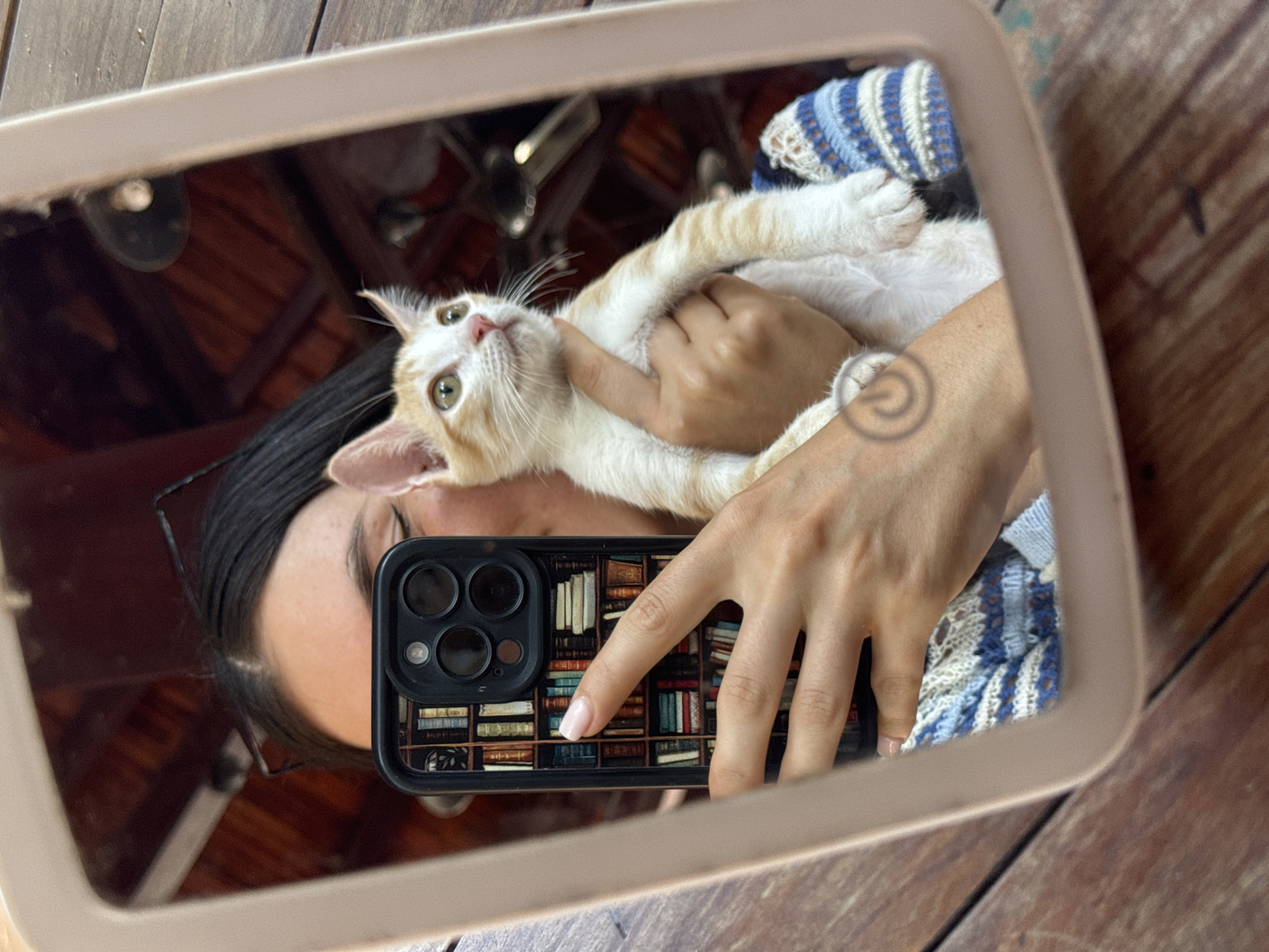 A person is taking a selfie in a mirror, holding a ginger and white kitten. The person's face is partially visible, and they are wearing glasses. The kitten is looking up and to the right. The person is holding a smartphone with a phone case designed to look like a bookshelf. The background of the mirror reflection shows a wooden ceiling and some lighting fixtures, suggesting an indoor environment with natural wood elements. The overall impression is a casual moment captured in a reflective surface. The image does not provide any explicit details that would allow for the identification of the location within Mũi Né, Vietnam.