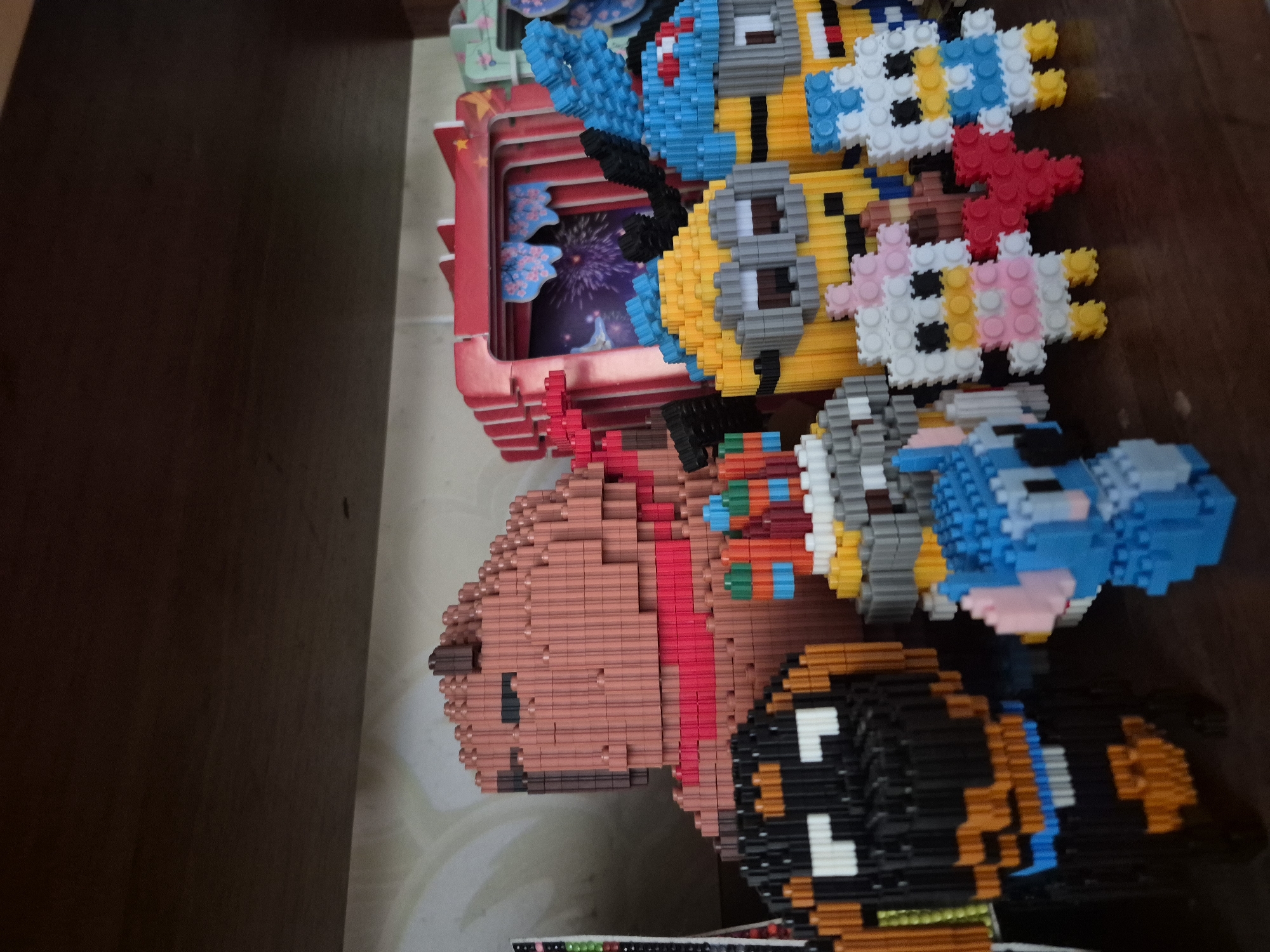 The image displays a collection of small, pixelated-style figurines, likely constructed from building blocks, arranged on a wooden shelf. These figurines include several characters that resemble Minions from the Despicable Me franchise, a blue character resembling Stitch from Lilo & Stitch, two duck-like figures, and a larger, brown, bear-like figure wearing a red scarf. Behind the Minion figures, a red, stage-like structure is visible with a backdrop depicting fireworks. The shelf itself appears to be made of dark wood, and the background behind the shelf is a light-colored, possibly textured wall. There are no people present in the image, and no actions or interactions are depicted. The setting is an indoor environment, likely a display or collection area. There is no information within the image to suggest a location in Kharnavy, Russia.