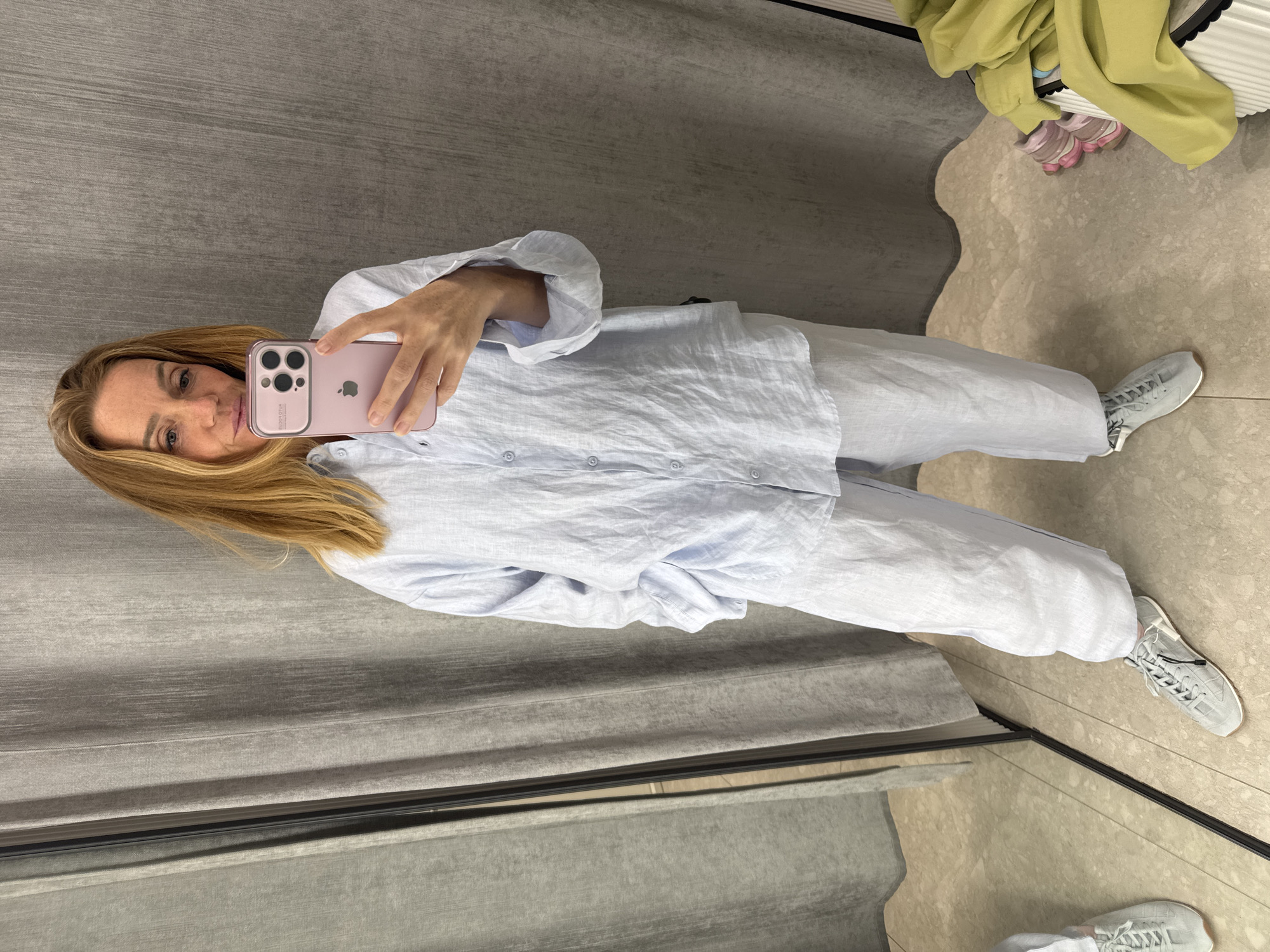 A woman with long, blonde hair takes a mirror selfie in what appears to be a retail changing room, located in Saint Petersburg, Russia. She is dressed in a light blue, long-sleeved linen button-up shirt and matching light blue wide-leg linen pants, creating a relaxed and coordinated outfit. On her feet, she wears light grey sneakers with white soles and dark grey laces.
She is holding a pink iPhone with a distinctive triple-lens camera system and the Apple logo visible on its back. The phone is encased in a rose-pink protective cover that prominently displays the text "LUXURY EDITION" and a Chinese character (光荣) near the bottom. Her face, reflected in the mirror, shows a neutral to slightly smiling expression.
The setting features grey, textured walls and a light, speckled tiled floor. A full-length mirror, framed in thin black, reflects the woman and portions of the room. In the background, on the floor, there is a pile of folded light green clothing, possibly a sweatshirt or hoodie, and a pair of small pink children's sneakers, suggesting additional items or companions. The activity captured is that of an individual trying on clothing and photographing the outfit.