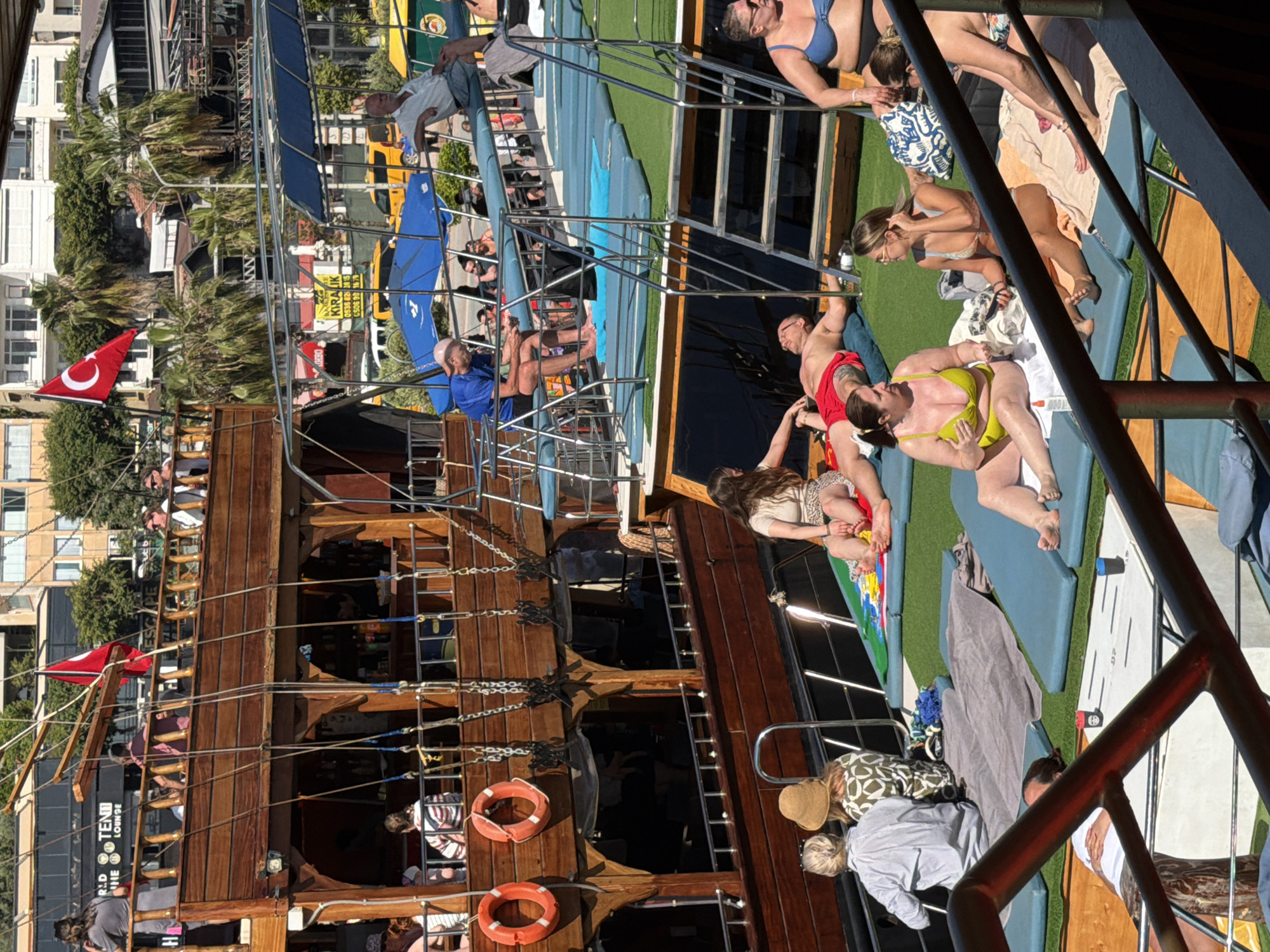 This image captures a vibrant daytime scene aboard a multi-decked wooden boat, likely an excursion vessel, docked or navigating in a coastal area of Alanya, Turkey. The weather appears sunny and clear, conducive to outdoor leisure.
The boat itself is a traditional-style wooden vessel with several levels, connected by metal ladders and railings. Its hull is dark wood, and decorative elements suggest a historical or rustic aesthetic. Two red life preservers are visible on the lower deck. A prominent Turkish flag (red with a white star and crescent) flies from a mast on the boat's port side, firmly establishing the location.
Numerous people are seen across the boat's decks, primarily adults enjoying the sunny weather. On the uppermost deck, individuals are sunbathing and relaxing on blue mats or towels. A woman in a blue bikini and sunglasses, another in a bright yellow bikini, and a man in red swim shorts are among those prominent here, appearing relaxed. Other passengers are seated or standing on the mid and lower decks, some observing the surroundings. The overall atmosphere suggests a leisurely and enjoyable tourist activity.
In the background, beyond the boat, a bustling shoreline is visible. Residential or hotel buildings, white and light-colored, line the coast. Palm trees are scattered throughout the landscape, indicative of a warm climate. On what appears to be a road or promenade behind the boat, yellow vehicles, possibly taxis, are parked or moving.
Visible text includes a partially obscured sign on the shore with "KİRALIK" (Turkish for "FOR RENT") and phone numbers (0538... and 0505...). Another banner in the background shows "WORLD TEN1 LOUNGE" and "RED ONE".