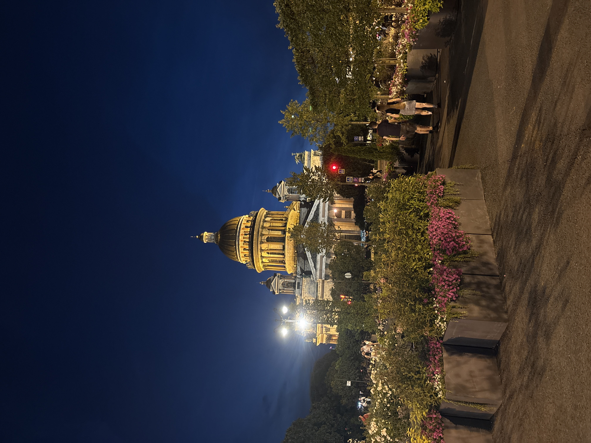 The image captures a night scene in Saint Petersburg, Russia, featuring Saint Isaac's Cathedral. The cathedral's golden dome and illuminated facade are prominent against a dark blue sky. In the foreground, a pathway is lined with large planters filled with lush green foliage and vibrant pink and white flowers. Two individuals, a man and a woman, walk away from the camera along the pathway. They are casually dressed, with the man wearing a dark t-shirt and shorts, and the woman a dark top and a skirt. In the background, more trees and planters are visible, along with streetlights and other figures of people in the distance, suggesting a public space or park. The overall atmosphere is serene and illuminated by artificial lights.