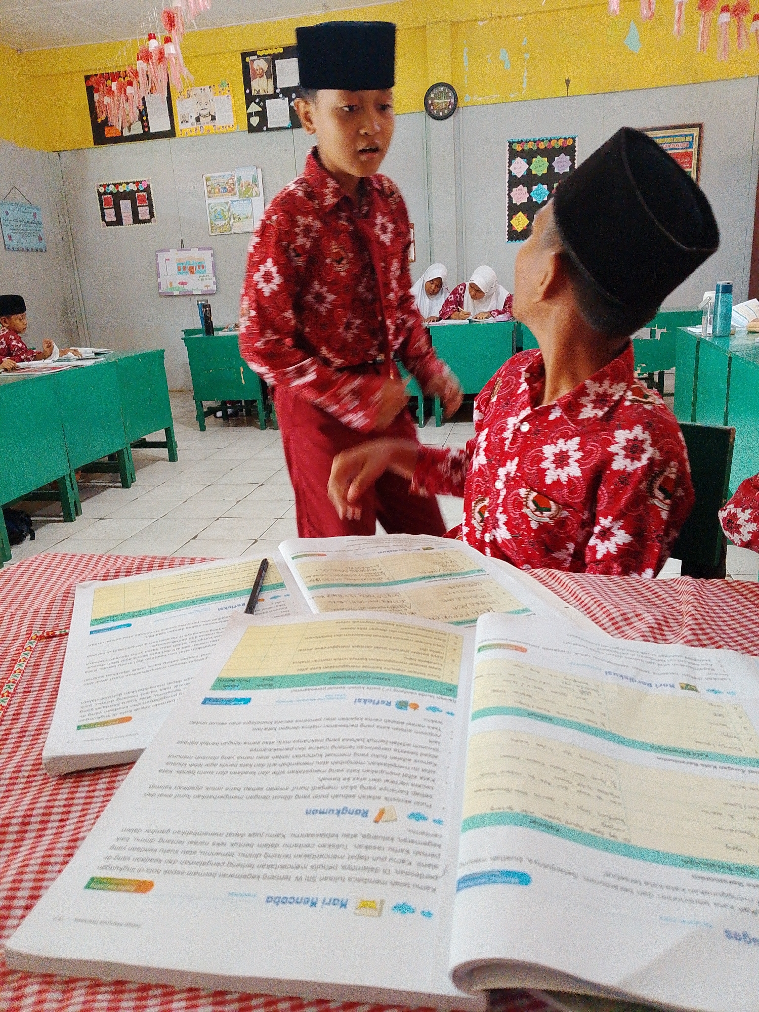 The image depicts an indoor scene, likely a classroom, with several young students. Two boys are prominently featured in the foreground and midground, both wearing red shirts with a white floral pattern and black peci hats. The boy in the foreground is facing away from the camera and towards the right. The boy in the midground is facing the camera and appears to be speaking or gesturing. In the background, two girls wearing white headscarves and similar red shirts are seated at desks. The classroom environment includes green desks, a yellow painted wall, and educational posters or decorations displayed on the walls. Books are open on a table covered with a red and white checkered tablecloth in the foreground. The presence of a clock on the wall and the style of the classroom suggest a school setting. There are no immediate indications pointing to the specific location of Tembung, Indonesia, beyond the implication that the image was taken there.