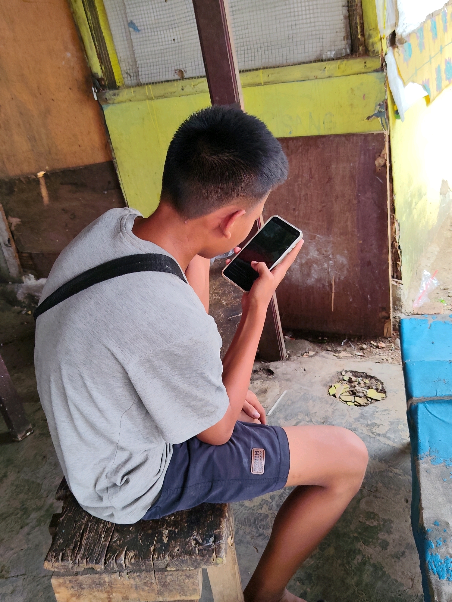 A young person, seen from behind and slightly to the side, sits on a rough wooden stool in an outdoor, unpaved area that appears to be an urban or semi-urban setting. The person is wearing a grey t-shirt with a black strap across the left shoulder and dark blue shorts. They are holding a smartphone in their right hand and appear to be looking at the screen, with their left index finger extended towards it.
The background is dilapidated, with walls of various materials including wood and yellow-painted plaster. One section of the wall is covered in a wire mesh, suggesting a partially enclosed structure. There is debris on the ground, including what appears to be broken tiles or concrete in a circular hole in the ground. A bright blue, weathered structure, possibly a bench or table, is visible to the right. The overall impression is one of a neglected or informal outdoor space. The scene is consistent with observations from Purwakarta, Indonesia, where similar visually deteriorated and informal structures are common.