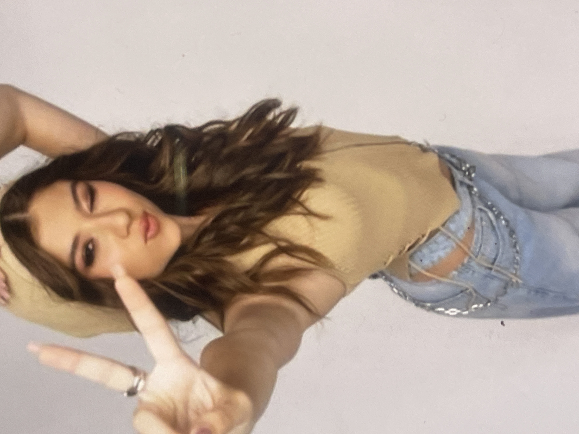 A portrait image, rotated 90 degrees clockwise, captures a young woman posing in what appears to be an indoor studio with a plain white background. The woman has long, wavy dark brown hair and is looking towards the camera with a playful expression, her lips slightly parted as if pouting.
She is wearing a light brown, possibly ribbed, sleeveless crop top that features lace-up details on the side, revealing a glimpse of her skin. Her attire also includes light blue jeans distinguished by large, intricate cut-outs on the hips, which are laced together and decorated with metallic silver-colored chains. Her left arm is extended forward, with her hand forming a peace sign and a prominent silver-toned ring visible on her index finger. Her right arm is raised and bent, with her hand positioned near her head. The lighting is soft and even, suggesting a controlled studio environment. No text is visible in the image. The provided context states this media file is from Poltava, Ukraine.
