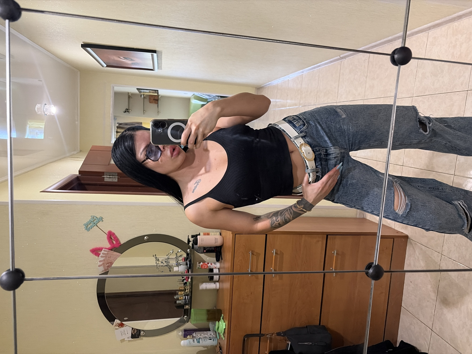 A woman is taking a mirror selfie in a room. She is wearing a black tank top, ripped blue jeans, and a white belt. She has long black hair and is wearing sunglasses. Her left arm, adorned with tattoos, is raised to hold her phone, which is angled to capture her reflection. The background shows a tiled floor, wooden cabinets, and a mirror. In the reflection of the mirror, there's a glimpse of a painting on the wall and what appears to be a light fixture. A small round mirror on the wall reflects more items, including pink bunny ears and some currency, possibly Russian Rubles given the location context of Ufa, Russia. The overall impression is a casual moment captured at home.