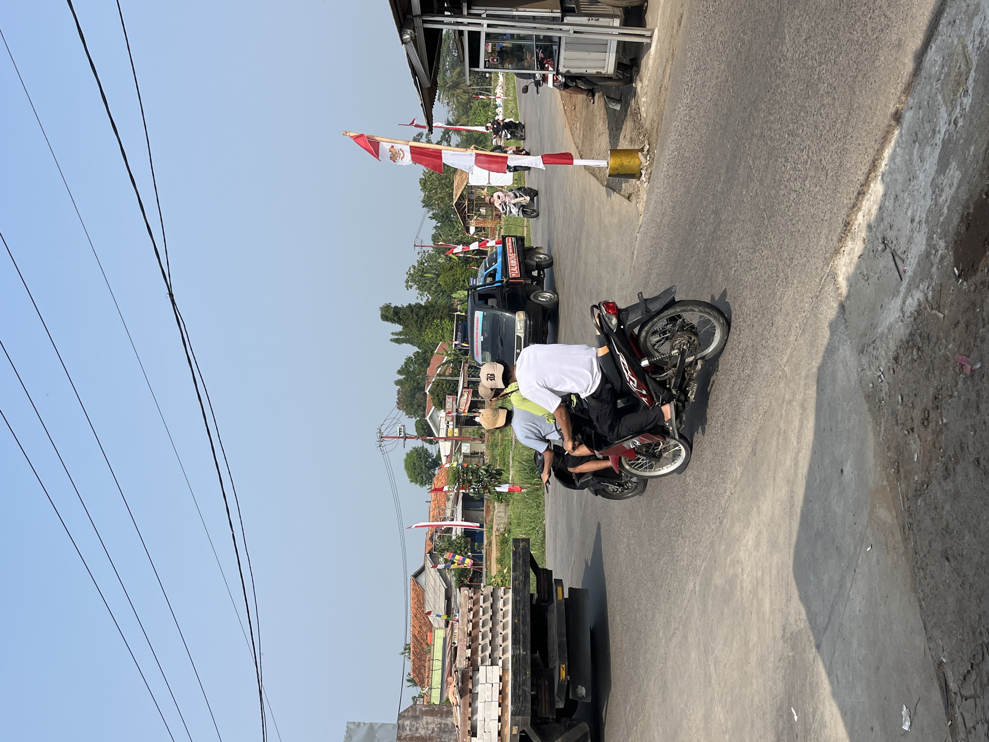 The image depicts a street scene in Bojong Gede, Indonesia, under a clear blue sky. In the foreground, two individuals are riding on a red motorcycle. The rider in front wears a white shirt and dark pants, while the passenger behind him wears a light blue shirt and dark pants. They are traveling on an asphalt road. To the left, a flatbed truck is parked, laden with stacks of bricks or tiles.
Further down the road, a black pickup truck with "Y LALAMOVE" written on its side is visible. Several Indonesian flags, red and white, are displayed along the roadside, tied to poles or attached to buildings. These flags suggest a festive occasion or a national celebration. A small roadside stall or structure with a white metal frame and a brown roof is situated on the right side of the road, with a motorcycle parked in front of it. In the background, residential buildings with red tiled roofs and scattered trees are visible. The overall environment appears to be a semi-urban or rural setting.