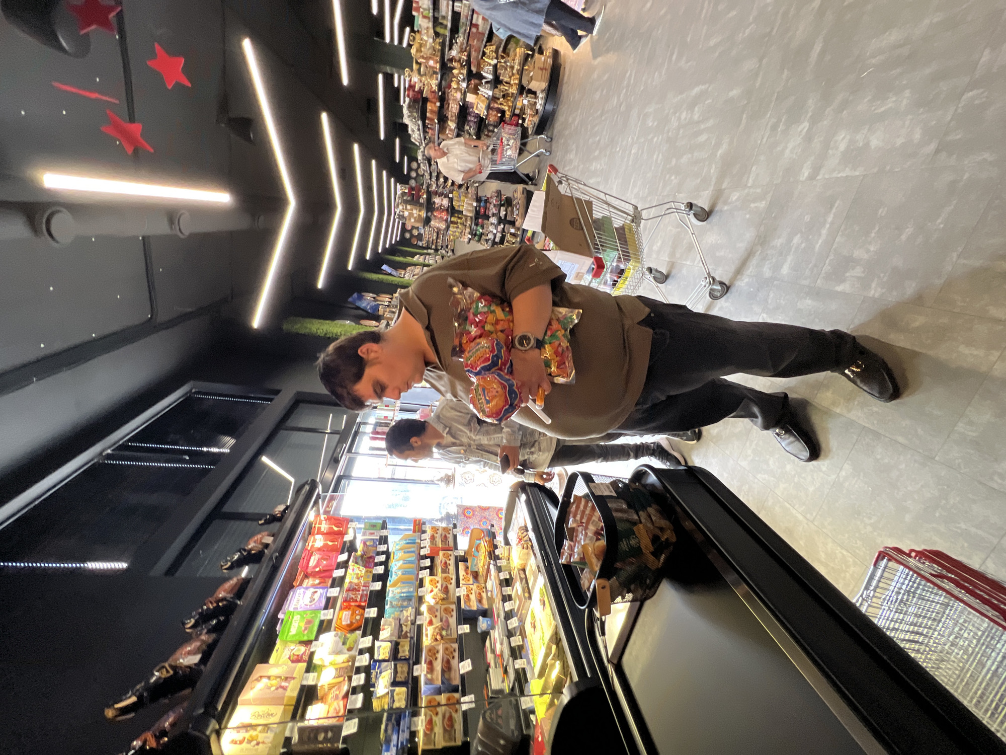 The image captures the interior of a modern supermarket or convenience store in Krasnodar, Russia, presented with a rotated perspective. The main subject, appearing to be male, stands prominently in the foreground, wearing a green/khaki short-sleeved shirt and dark pants. They are holding two clear bags filled with colorful candies or gummies and are looking downwards, likely at products on a shelf or display. A watch is visible on their left wrist.
Behind the primary subject, another person in a light-colored top is partially visible, also engaged in browsing. Further down the aisle, a person in a white shirt is seen pushing a shopping cart. The store's interior features dark walls and ceilings decorated with striking linear LED lights arranged in arrow-like patterns, complemented by red star ornaments and small white dots. The floor is a light grey, possibly tiled or concrete.
Shelves line the aisles, stocked with various packaged goods, some appearing to be bottles or snack items. Black display cases are visible in the foreground. Several shopping carts are scattered throughout the store, one containing a cardboard box, and a stack of red carts is visible in the lower right corner. The overall activity involves customers shopping and browsing the merchandise. The store is brightly illuminated by artificial lighting. No discernible text is clearly visible on products or signs within the image.