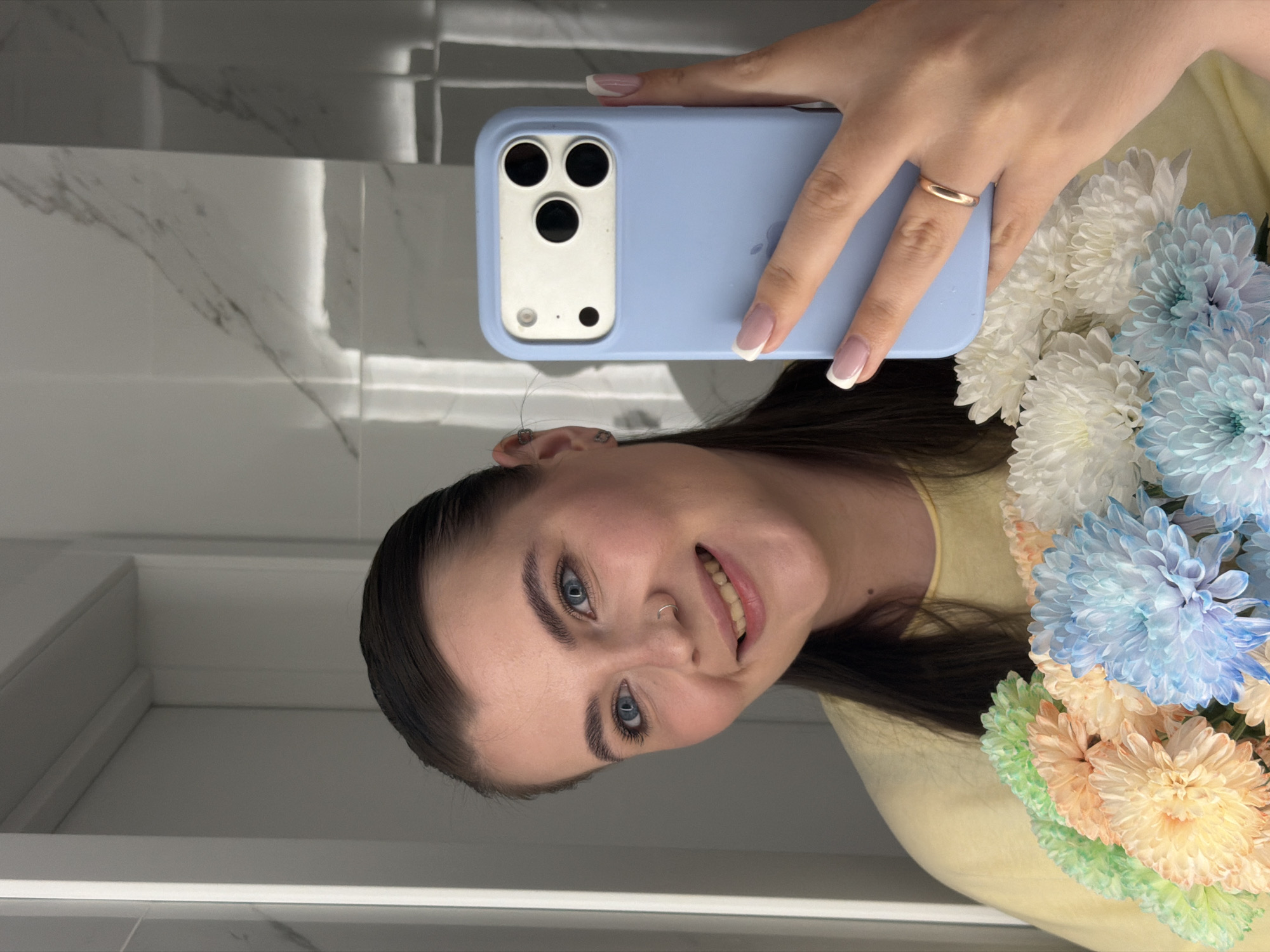 The vertical image presents a mirror selfie of a young woman in an indoor setting. She has dark hair, blue eyes, and a nose piercing, and she wears a gentle, contented smile. Her attire includes a light yellow top.
In her right hand, she holds a light blue smartphone, likely an iPhone given its distinct camera array with three lenses and a flash, encased in a matching light blue cover. Her fingernails are manicured with a classic French tip style, and a simple gold band ring adorns her right ring finger. Small silver stud earrings are visible in her left ear.
Alongside the phone, she holds a colorful bouquet of flowers, resembling chrysanthemums. The blossoms feature a soft palette of white, pastel blue, light orange/peach, and light green.
The background reflected in the mirror shows a clean, modern interior, likely a bathroom, characterized by white tiled walls with subtle grey marble veining. A white counter or ledge is also visible. The bright, artificial lighting suggests an indoor environment. The Apple logo is faintly visible on the back of the phone case. The image does not provide cues for the time of day or outdoor weather. This scene is set in Chaikovsky, Russia.