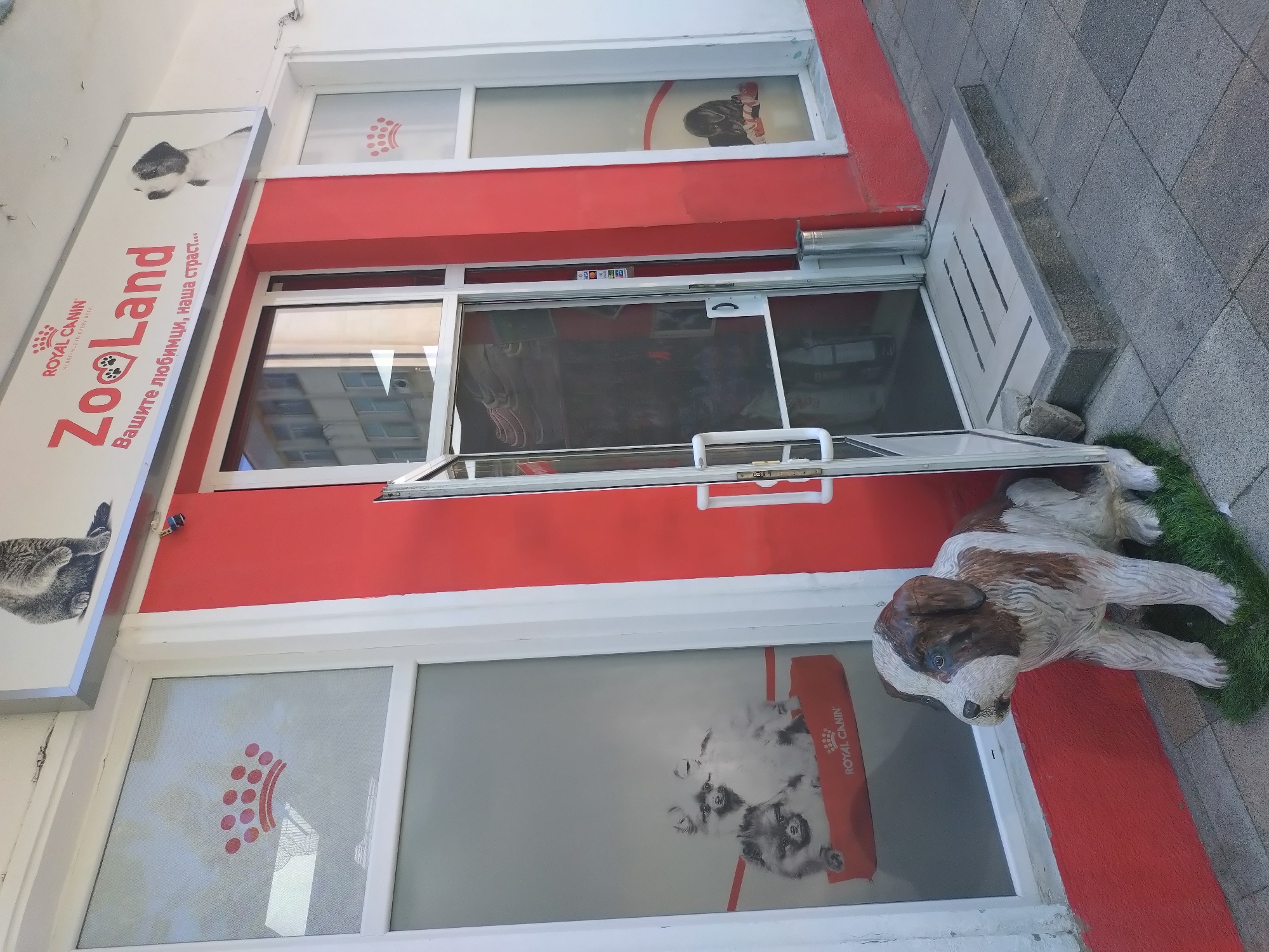 The image shows the storefront of a pet supply store called "Zoo Land" located in Stara Zagora, Bulgaria. The store's sign displays the "Royal Canin" logo. The storefront is painted in red and white, with large window displays. One window display features a graphic of cats, while another shows a graphic of dogs. There is also a statue of a dog near the entrance to the store. The entrance is a glass door that is partially open.