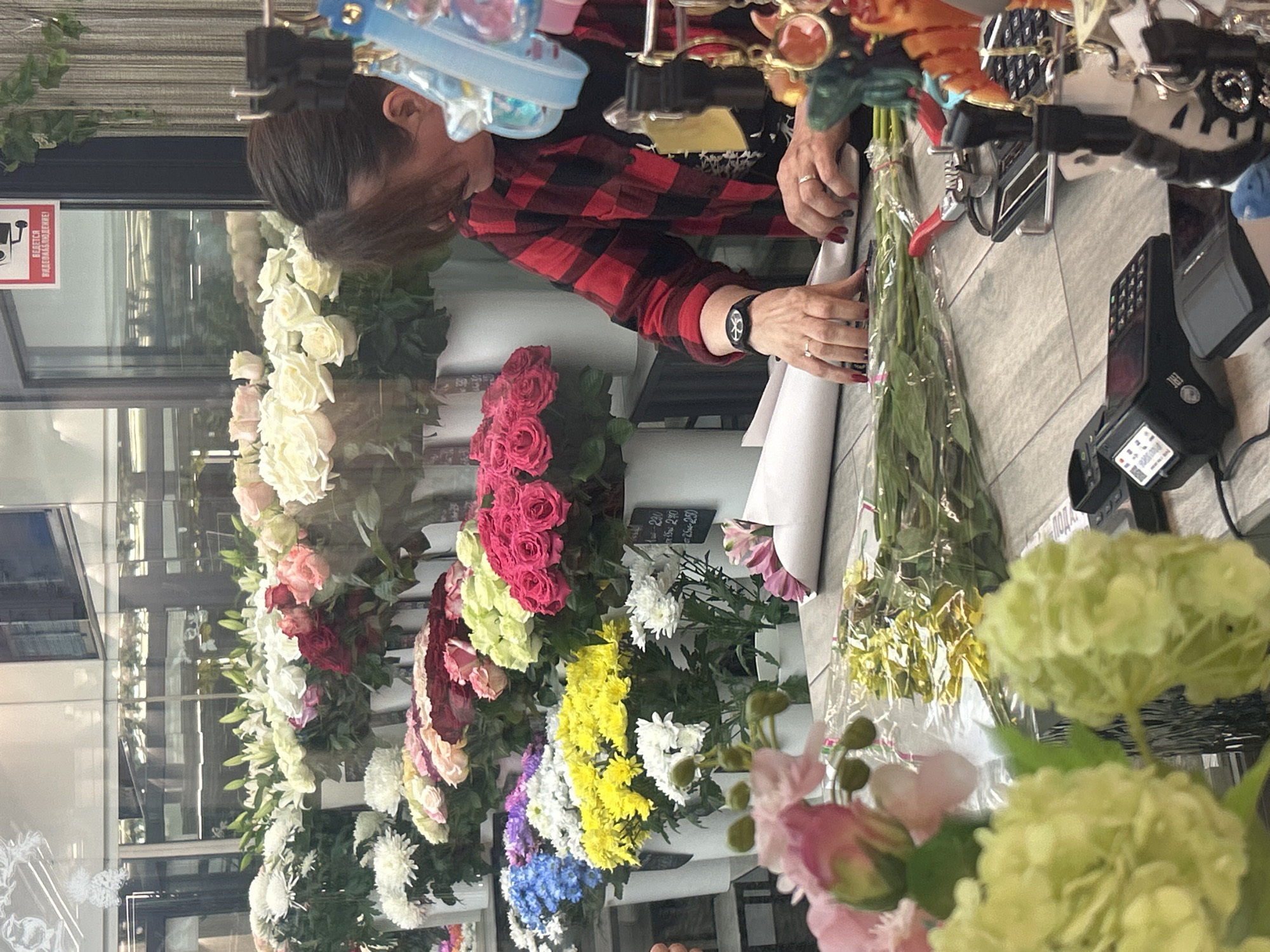 The image captures a close-up view of a flower shop counter in Saint Petersburg, Russia, focusing on a woman arranging a bouquet. The woman, with dark hair pulled back, wears a red and black plaid shirt. Her hands, adorned with rings and dark red nail polish, are actively working with a bouquet wrapped in white paper and green stems. To her right, a pair of pruning shears rests on the counter. Several payment terminals and a calculator are visible on the counter, suggesting a retail environment. The background is filled with a vibrant display of various flowers in white buckets, including white chrysanthemums, roses in shades of pink, red, and yellow, and blue hydrangeas. The overall impression is that of a busy flower shop on a potentially sunny day, given the bright lighting.