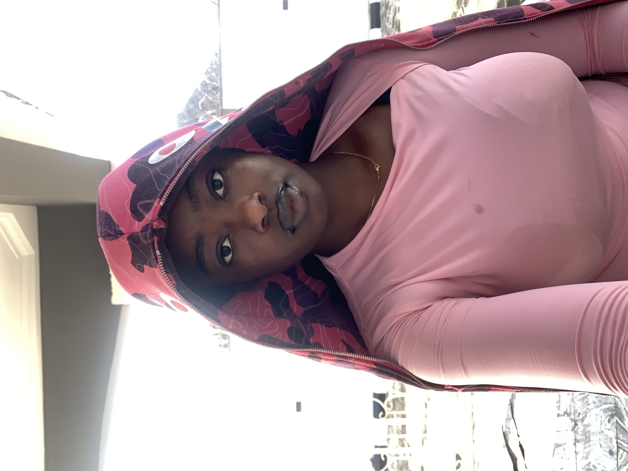 A young Black woman with dark skin and dark hair is shown in a close-up, waist-up selfie, looking directly at the camera. She is wearing a pink long-sleeved shirt and an open, hooded jacket with a pink and purple camouflage pattern. A thin gold chain with a small pendant is visible around her neck. Her lips are dark and glossy, and her tongue is slightly extended. The background appears to be outdoors with a white wall and some blurred elements that suggest a natural or urban setting, possibly in Gura Top, Nigeria. The lighting is bright, coming from the right side of the frame.