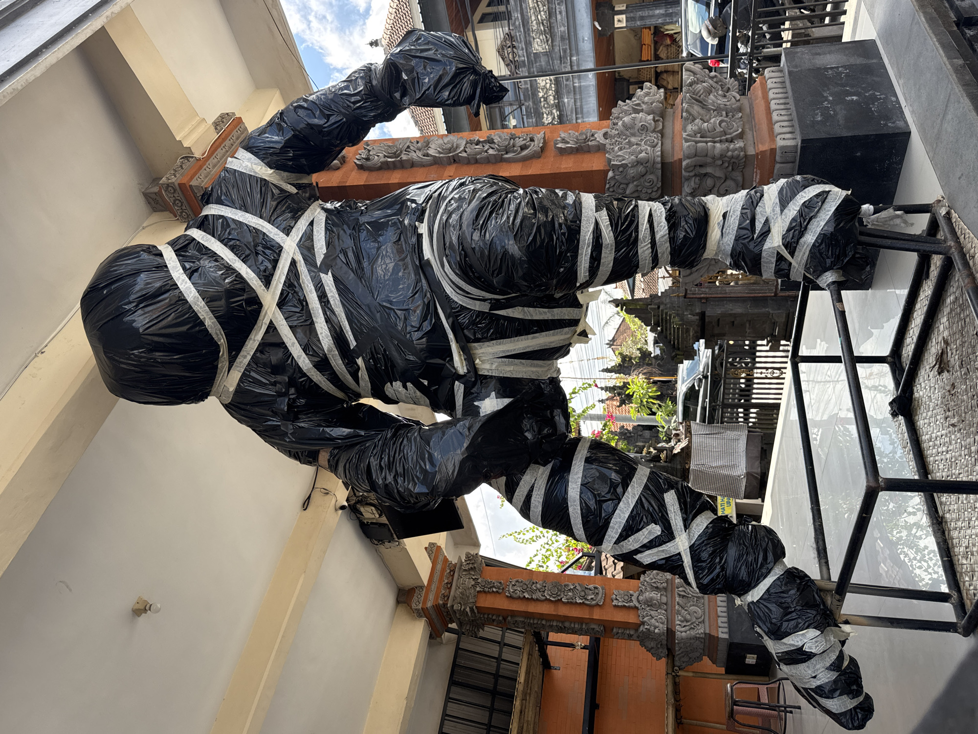 An imposing, life-sized sculpture, possibly a representation of a figure or creature, stands draped in black plastic sheeting and bound with white tape. The sculpture has a bulky, amorphous form, with its limbs wrapped and secured in a manner suggestive of mummification or restraint. It is positioned outdoors, under a covered walkway or balcony with cream-colored beams and a light-colored ceiling.
The sculpture's feet rest on a metal frame structure that appears to be part of its display. Behind the sculpture, ornate stone pillars with intricate carvings are visible, indicating a traditional architectural style. The background reveals elements of an urban environment, with buildings, fences, and vehicles, suggesting it is located in a populated area. Lush greenery and trees can be seen in the distance, hinting at a tropical climate. The sky is partially visible, with a few clouds present.
The scene is likely situated in the Ubud District of Indonesia, given the architectural style and the general ambiance. The overall impression is one of an artistic display or a temporary installation, possibly for a cultural event or festival. The lighting suggests it is daytime.
