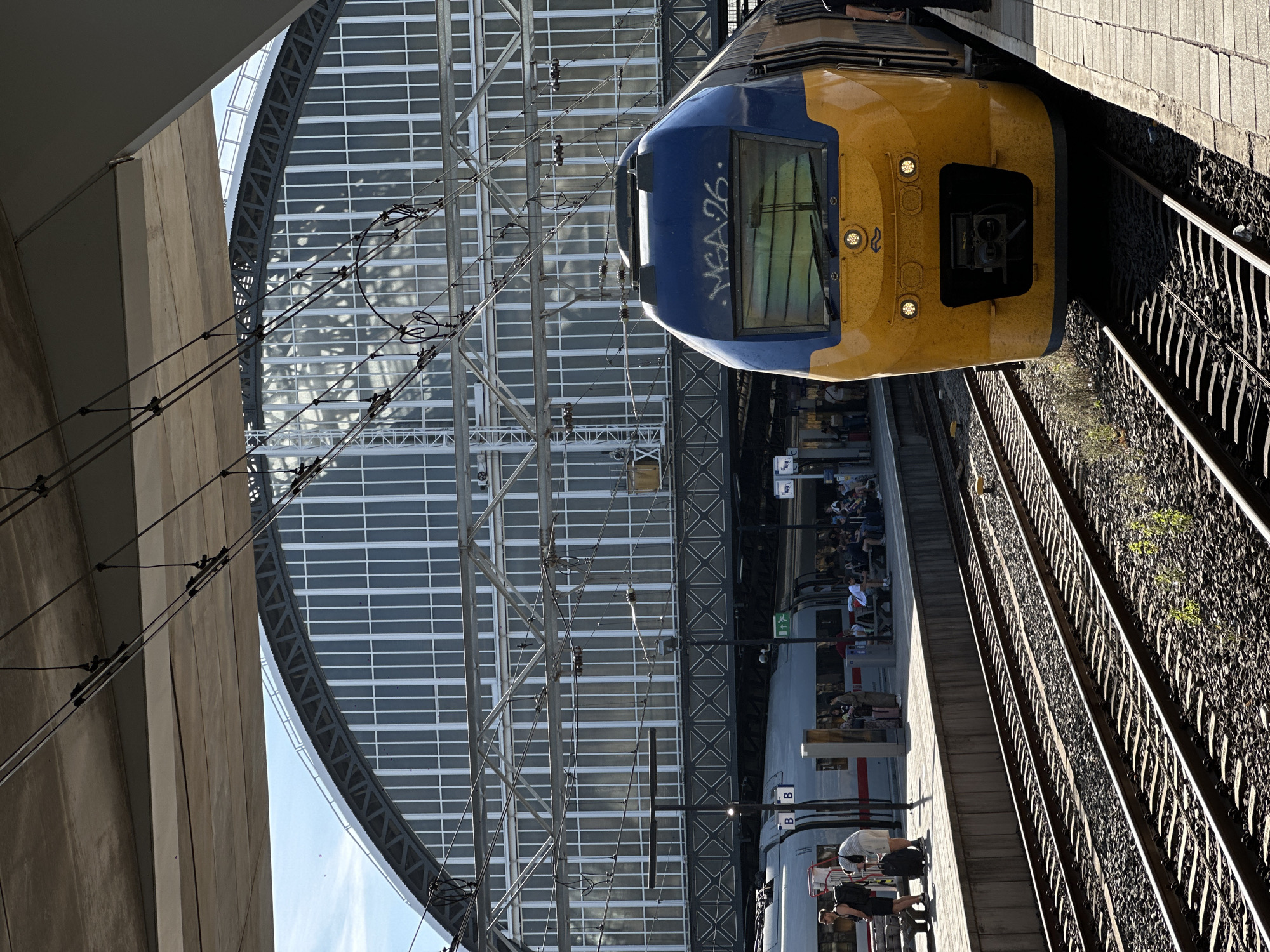 The image shows a view of Amsterdam Centraal Station, a major railway hub in Amsterdam, Netherlands. In the foreground, a yellow and blue train with the graffiti "MBA26" on its front is parked on the tracks. The train has its headlights on and a small symbol of a leaping deer is visible on its front. Further back, another train, primarily grey with red accents, is visible on an adjacent platform. People are standing and sitting on the platform, some with luggage, waiting or disembarking. Above the platforms, a large, arched glass and steel structure forms the roof of the station, with numerous electrical wires for the trains suspended beneath it. The sky is blue and the scene is bathed in sunlight, casting shadows. The environment is a bustling urban train station on a clear day.