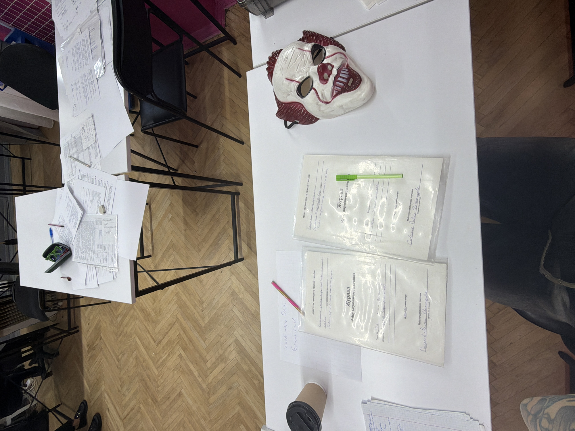 The image depicts a white table with various documents, a clown mask, a coffee cup, and pens. The documents appear to be official papers, some in plastic sleeves, with handwritten text. The clown mask is a disturbing, realistic mask with white and red makeup. A brown paper coffee cup with a black lid is on the left side of the table. Several pens, including a pink one and a green one, are also visible. In the background, there are folding chairs and a wooden floor. The scene appears to be set up for some kind of event or registration process.