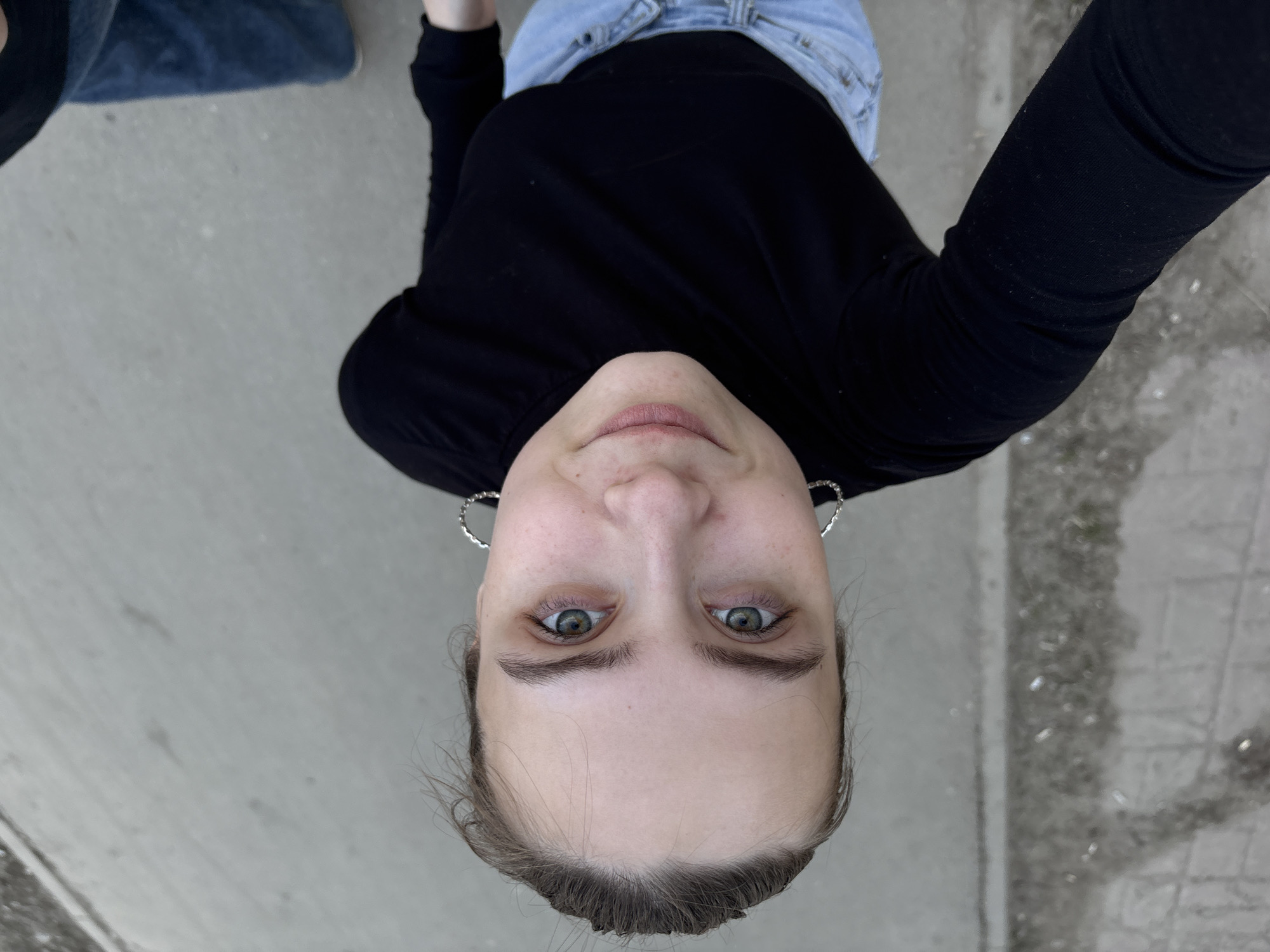 An overhead selfie captures a young person looking directly up at the camera, taken from a very low angle. The subject, a person with fair skin, has their head tilted back, revealing wide, light-colored eyes and a subtle, closed-mouth smile. They wear a simple black long-sleeved top, and a portion of light blue denim jeans is visible around their waist at the top of the frame. Silver-colored hoop earrings are seen on each ear. Their hair appears to be pulled back, exposing their forehead. The background consists entirely of a flat, light gray concrete or paved surface, which forms the ground beneath the subject. Some areas of the ground exhibit darker patches or variations in texture, hinting at an outdoor setting, possibly a street or pedestrian area. The lighting suggests it is daytime, with soft, indirect illumination. The activity is clearly a self-portrait, with the camera positioned below the subject's face. No text is visible in the image. The photo depicts a scene in Votkinsk, Russia.