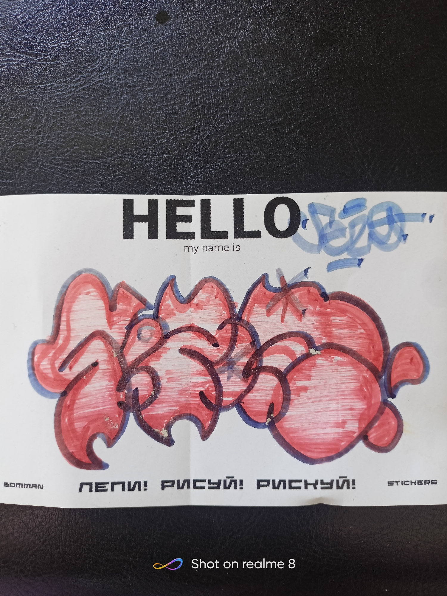The image shows a close-up, eye-level view of a piece of paper with drawings and text on it, placed on a black, textured surface, possibly a leather-bound book or a car seat. The paper has a white background and features a bold, black text reading "HELLO" on the upper left side. To the right of "HELLO," there is a smaller, handwritten phrase in blue marker that appears to be a stylized signature or tag. Below "HELLO," in smaller black font, is the text "my name is."
Dominating the lower portion of the paper is a colorful graffiti-style drawing rendered in red and outlined in dark blue and black. The drawing is abstract, with flowing, bubble-like shapes and sharp, angular elements, suggesting a stylized word or character.
Along the bottom edge of the paper, there are several text elements. On the left, a small logo or text reads "BOMMAN". In the center, in black block letters, are the Russian words "ПЕПИ! РИСУЙ! РИСКУЙ!", which translate to "Pepi! Draw! Risk!". On the right, in smaller black letters, is the word "STICKERS".
The image also contains a watermark in the bottom right corner that reads "Shot on realme 8" along with a colorful infinity symbol. The overall impression is of a creative piece, possibly a sticker design, presented in a casual setting. There is no indication of the city of Shelekhov, Russia, within the image itself.