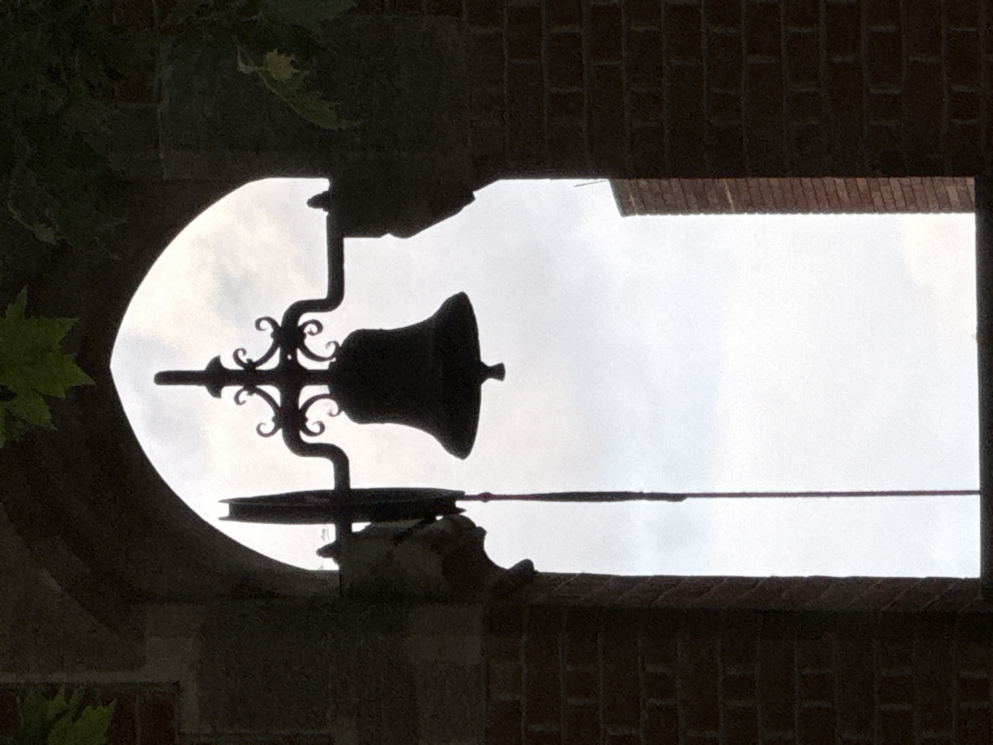 The image depicts a bell hanging in a structure with two openings against a cloudy sky. The bell is dark, with decorative ironwork. The structure appears to be made of brick and stone, with a round opening on the left and a rectangular one on the right. Vegetation is visible to the left of the image. The exact location is not determined, but the style suggests an old bell tower, potentially in London, United Kingdom. No people are visible.