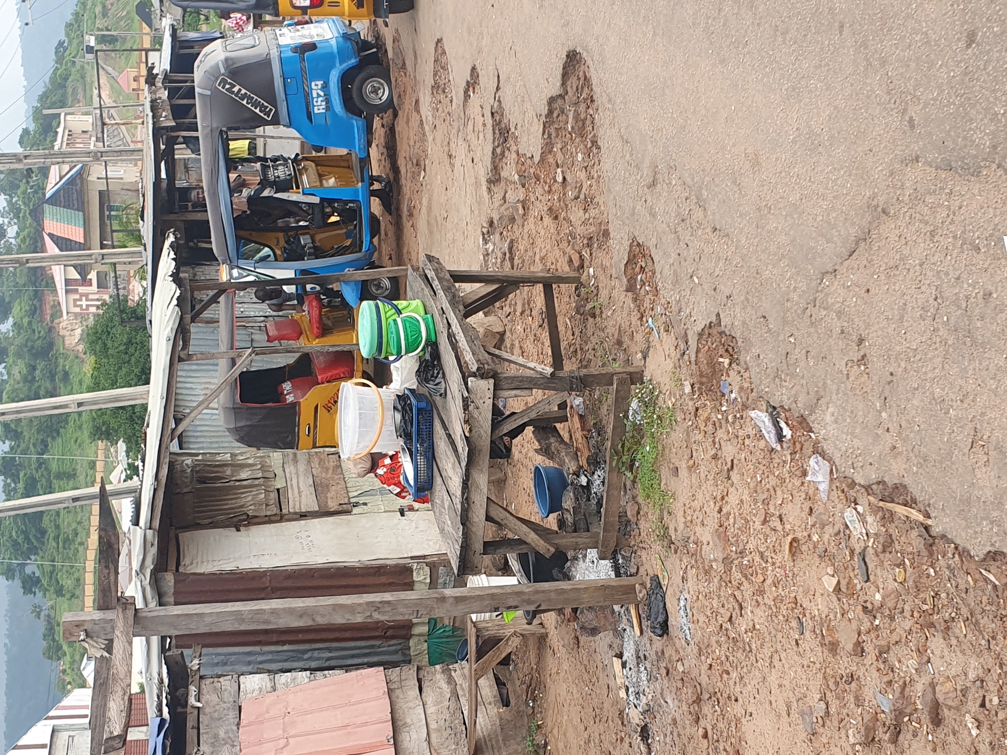The image depicts a street scene in Lokoja, Nigeria, characterized by a rustic and somewhat unkempt environment. A prominent feature is a roadside stall constructed from makeshift materials, including corrugated metal and wood. In front of this stall, a wooden table displays a blue plastic basket containing various items, possibly food or goods for sale, alongside two buckets. One bucket is green and the other is white.
Several auto-rickshaws, commonly known as Keke Napep, are visible. A blue one is parked in the foreground, with its door open. A yellow Keke Napep is partially visible to its left, and another to the right. The road surface in front of the stall is deteriorated, with several large potholes and scattered debris, indicating poor road conditions.
The background reveals a hilly landscape with vegetation and some buildings, including a structure with a distinctive multi-colored roof and a cross, suggesting it might be a church. Power lines and poles are also present, suggesting access to electricity. The overall atmosphere is that of a typical, bustling roadside market area in a Nigerian city, with an emphasis on functional, temporary structures and everyday commerce.