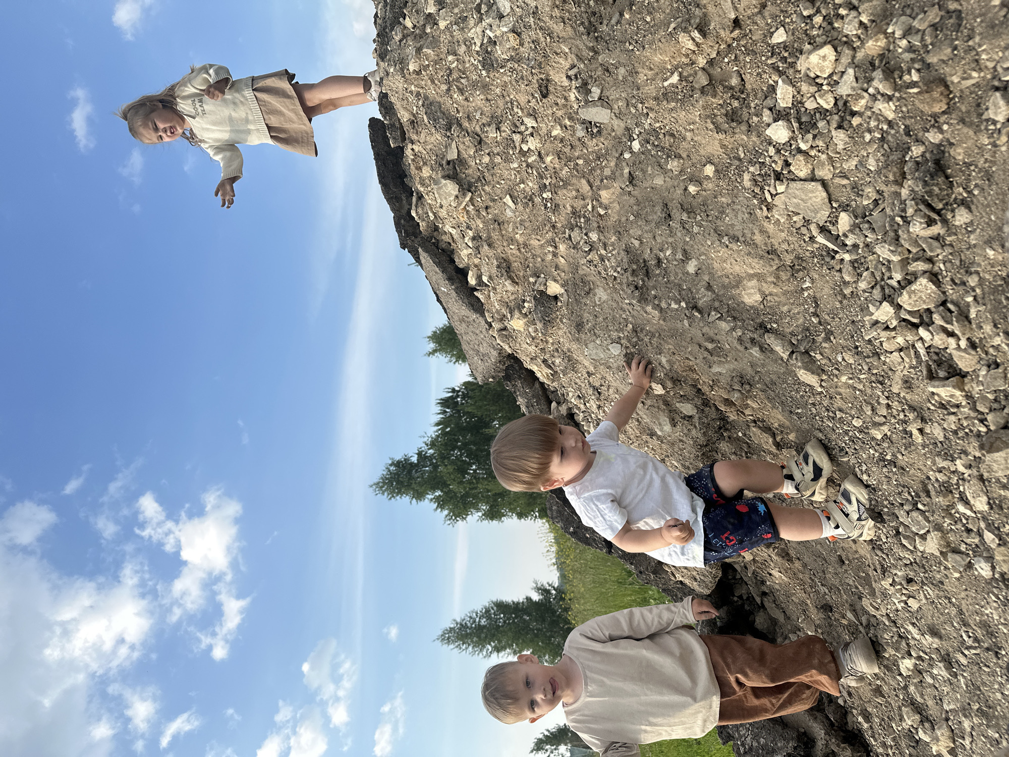Three young children are interacting on a dirt and asphalt mound under a clear blue sky with scattered white clouds. In the foreground, a boy in a white t-shirt and patterned shorts is climbing the mound, reaching out with his right hand. To his left and slightly behind him, another boy in a beige long-sleeved shirt and brown pants stands facing the camera, with his tongue sticking out slightly. In the background, on top of the mound, a girl in a cream-colored sweater and a tan skirt is looking to her left with her arms outstretched. The environment appears to be an outdoor, undeveloped area, possibly a construction or demolition site, with sparse trees and grassy fields visible in the distance. The location is identified as Estachi, Russia.