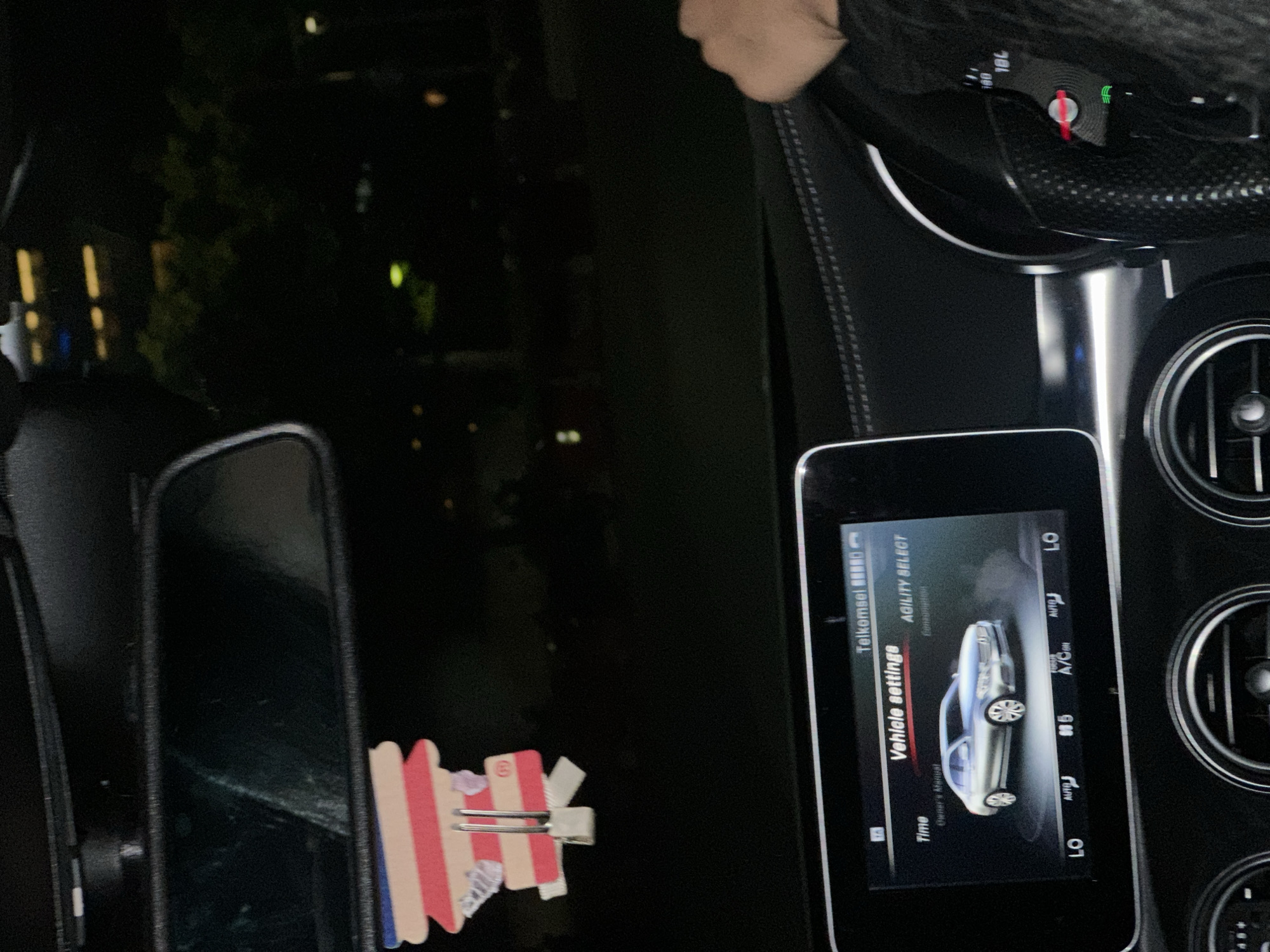 The image shows the interior of a car, likely a Mercedes-Benz, displaying the vehicle settings on the central display screen. The screen shows information like “Vehicle settings”, “Time”, “Owner’s Manual”, “Agility Select”, and “A/C ON”. The dashboard and air vents are visible, along with the steering wheel partially in the frame. An air freshener in the shape of a flag is seen attached near the rearview mirror. The lighting is dim, suggesting it is night. The location, as inferred from the text "Telkomsel" on the screen, is most likely in Indonesia. Central Jakarta, Indonesia is mentioned in the prompt.