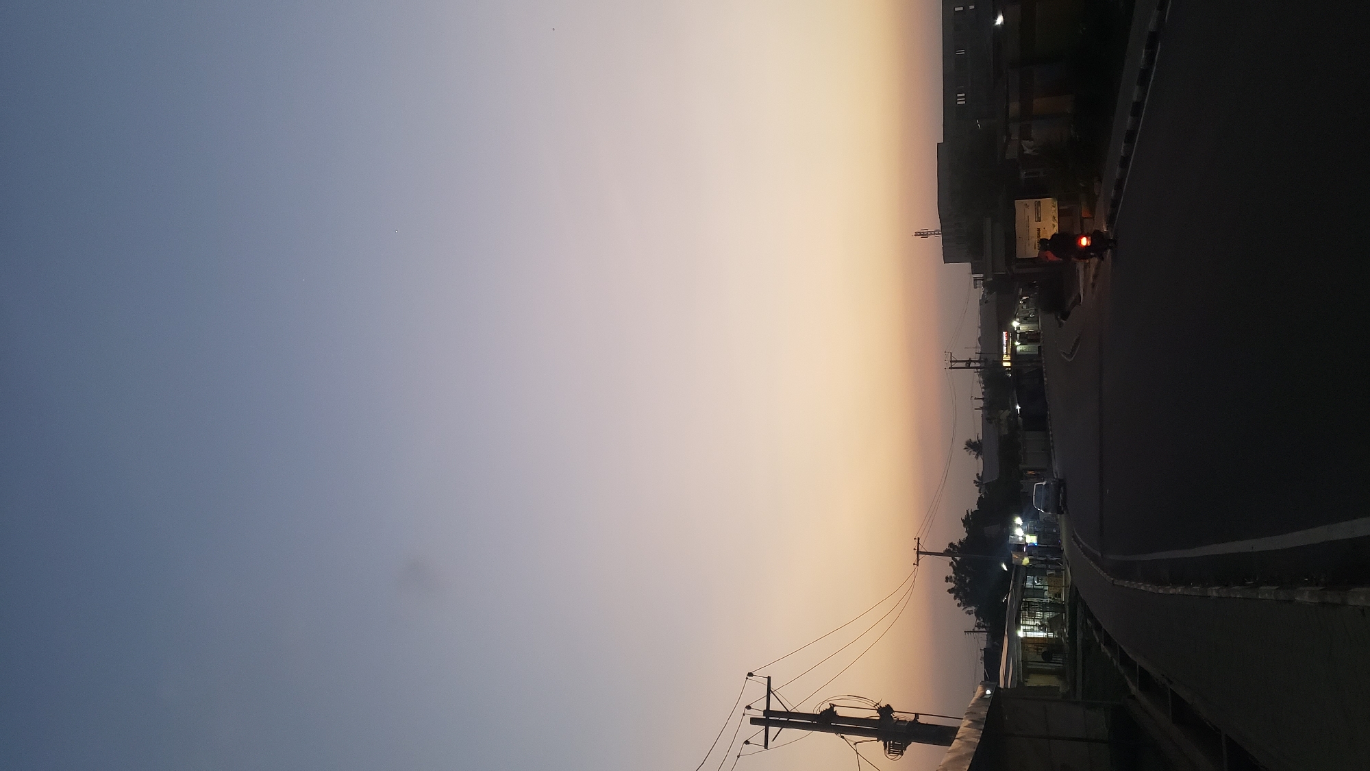 The image depicts an outdoor scene at either dusk or dawn, characterized by a striking gradient in the sky. The sky transitions from a deep blue-grey on the left to a warm, soft orange-yellow on the right, indicating the setting or rising sun. Faint specks of light, possibly stars or distant objects, are visible in the bluer part of the sky.
Below the horizon, an urban or semi-urban landscape unfolds. A dark asphalt road, likely a main thoroughfare, occupies the lower right portion of the frame, curving gently into the distance. A clear white line marks its edge. Several vehicles are visible on the road: a motorcycle with a distinctive glowing red tail light in the mid-ground, and at least one other dark car further down the road, appearing as a silhouette.
Along the left side of the road, various buildings are silhouetted against the fading light. Some buildings show illuminated windows or signs, casting a soft glow. Numerous utility poles with a network of power lines are prominent in the foreground and mid-ground, standing starkly against the colorful sky. Dark silhouettes of trees and other vegetation line the roadside.
The scene captures the tranquil transition between day and night, with artificial lights beginning to punctuate the fading natural light. The overall atmosphere suggests daily life continuing as darkness approaches or recedes. No discernible text is visible in the image. Given the context of Narayi, Nigeria, this image offers a glimpse into an evening or morning street scene in that region.