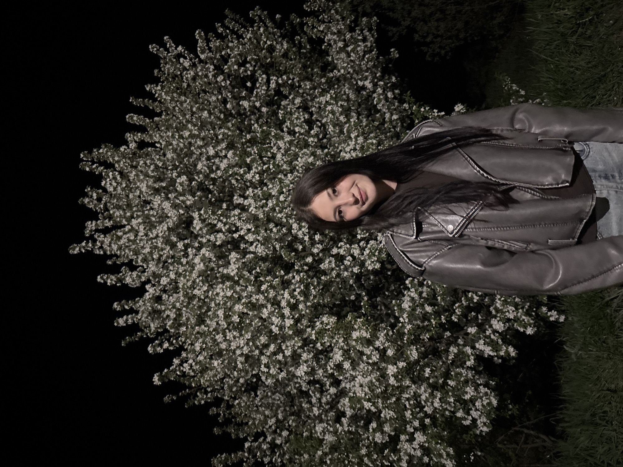 A young woman with long, dark hair stands near a blossoming tree at night in what appears to be a park setting. She wears a gray leather jacket and jeans. The tree is covered in small, white flowers, contrasting with the darkness of the night sky. The right side of the image suggests a grassy area. There is no indication of the location being Фоки, Russia.